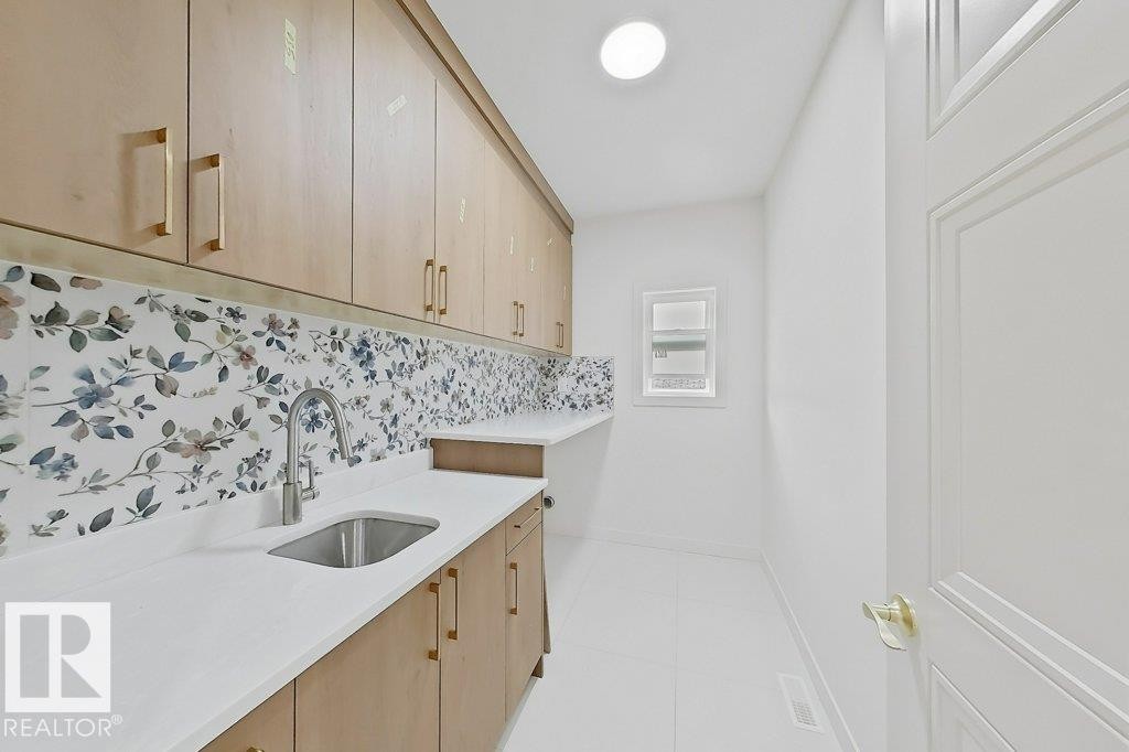 Light-filled utility space featuring extensive light wood-finish cabinetry with gold-tone hardware, a floral-patterned backsplash, white countertops, and an undermount sink with a gooseneck faucet - 9216 91 Street Nw, Edmonton, AB - Indoor Photo Showing Kitchen