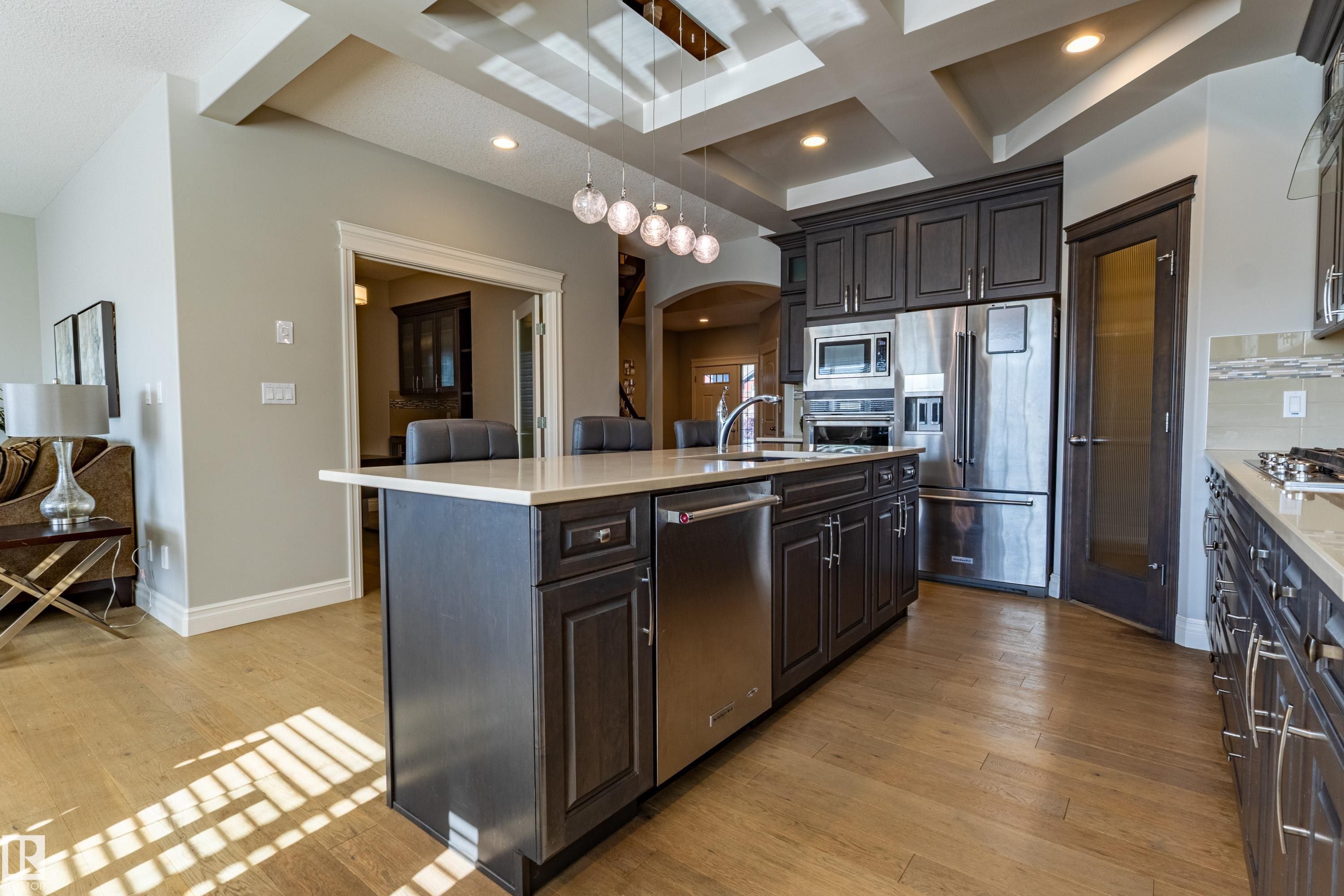Kitchen featuring dark wood cabinetry, stainless steel appliances, a large island with a light-toned countertop, wood-finish flooring, and a coffered ceiling with recessed lighting - 233 Hawks Ridge Boulevard Nw, Edmonton, AB