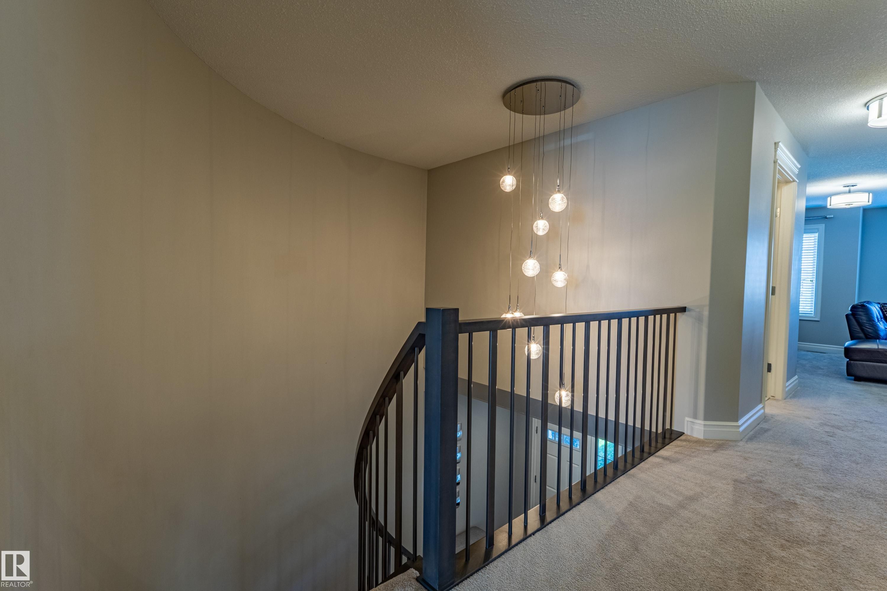 Curved staircase with black metal balusters, contemporary pendant light fixture, and carpeted hallway - 233 Hawks Ridge Boulevard Nw, Edmonton, AB