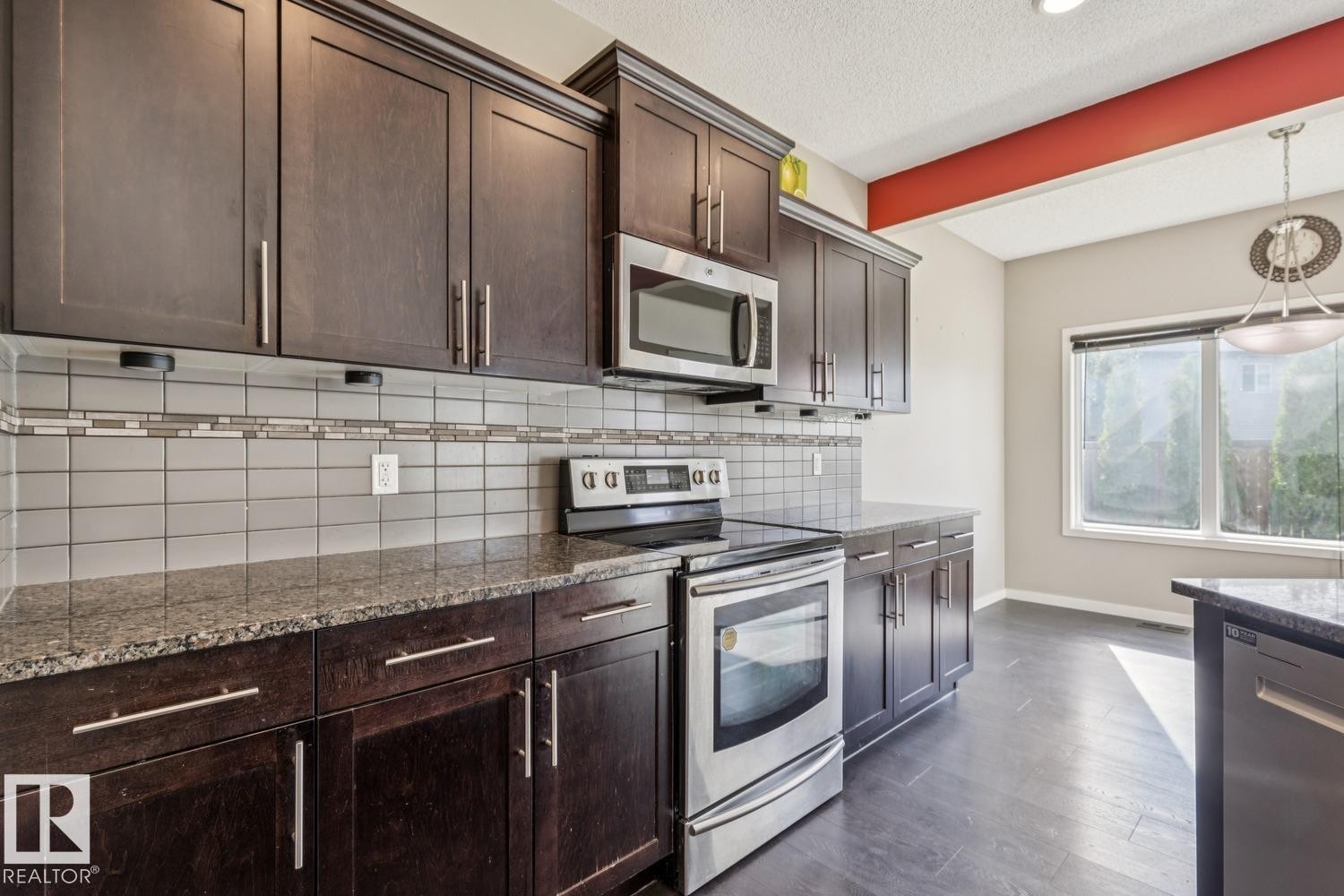 Kitchen featuring dark wood cabinetry, stainless steel appliances, speckled countertops, and a tile backsplash with decorative accent banding - 12007 173 Avenue Nw, Edmonton, AB