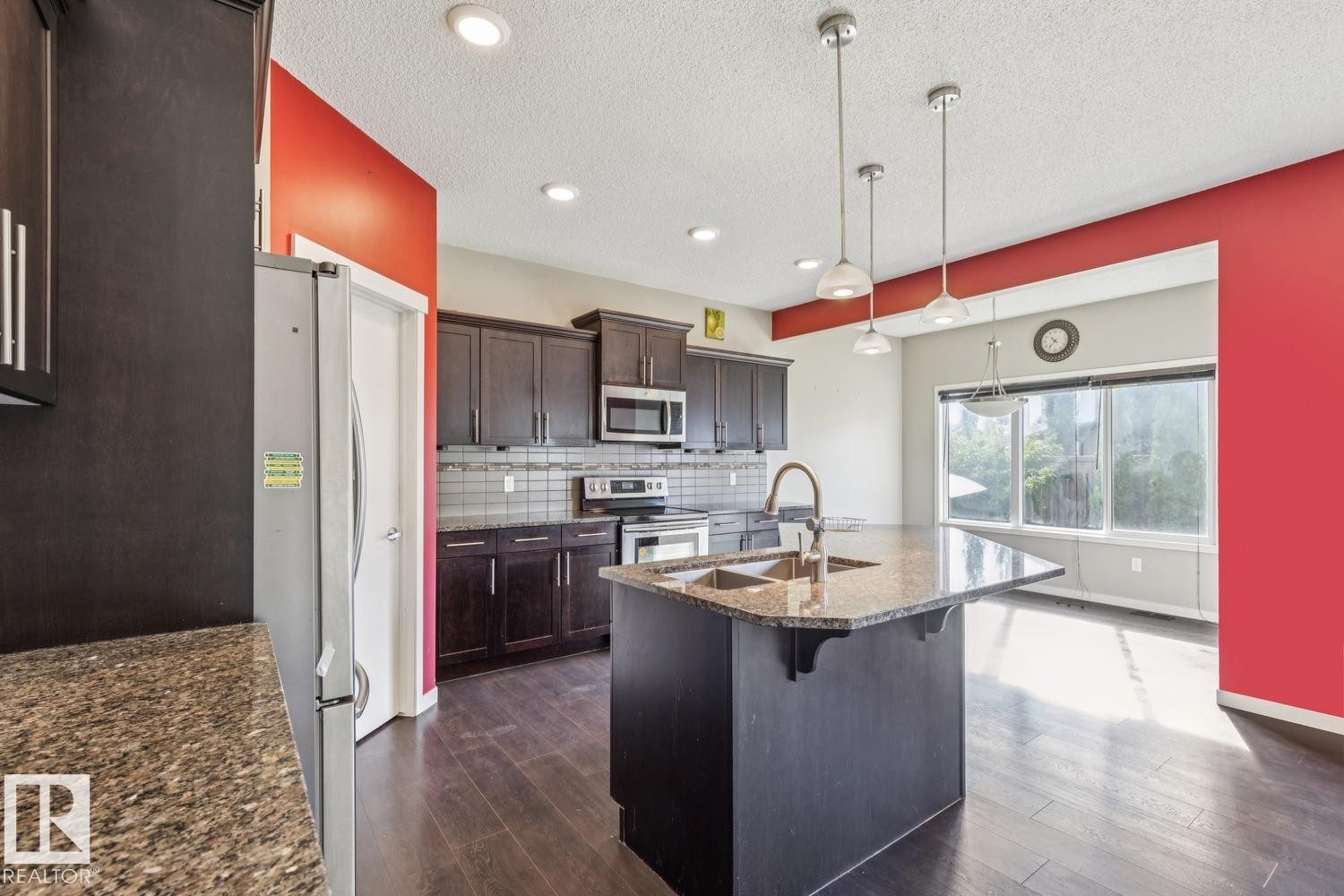 Kitchen featuring dark wood-finish cabinetry, stainless steel appliances, speckled stone countertops, a central island with an undermount sink, and recessed lighting - 12007 173 Avenue Nw, Edmonton, AB