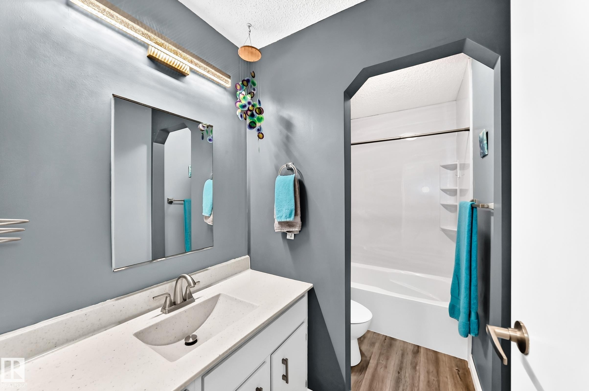Vanity with integrated sink, modern wall-mounted light fixture, full-width mirror, and a shower-tub combination with wood-finish flooring - 5620 93A Avenue Nw, Edmonton, AB