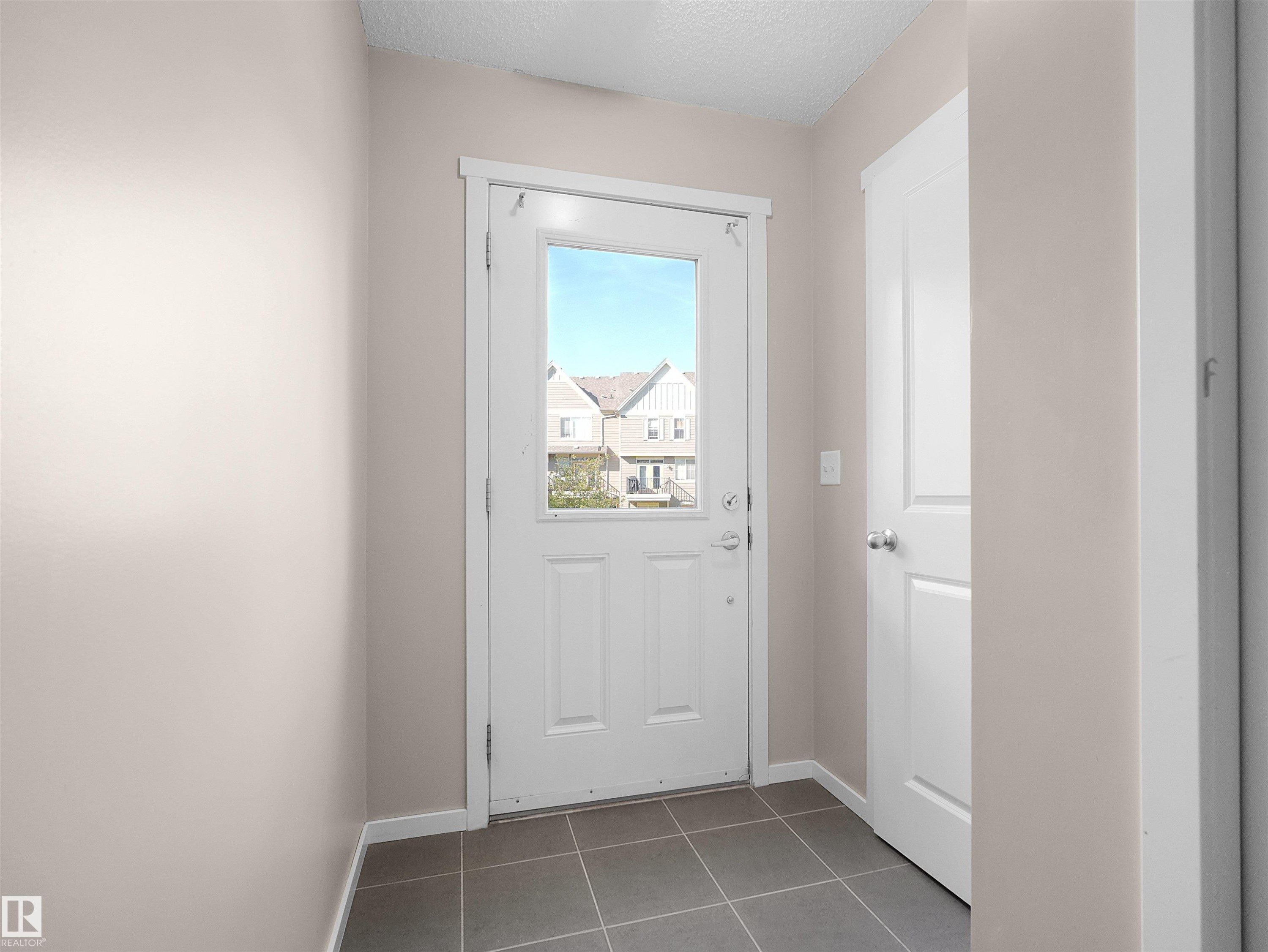 Entryway featuring a white paneled door with an integrated window, dark gray square tile flooring, and light neutral painted walls - #162 603 Watt Bv Sw, Edmonton, AB