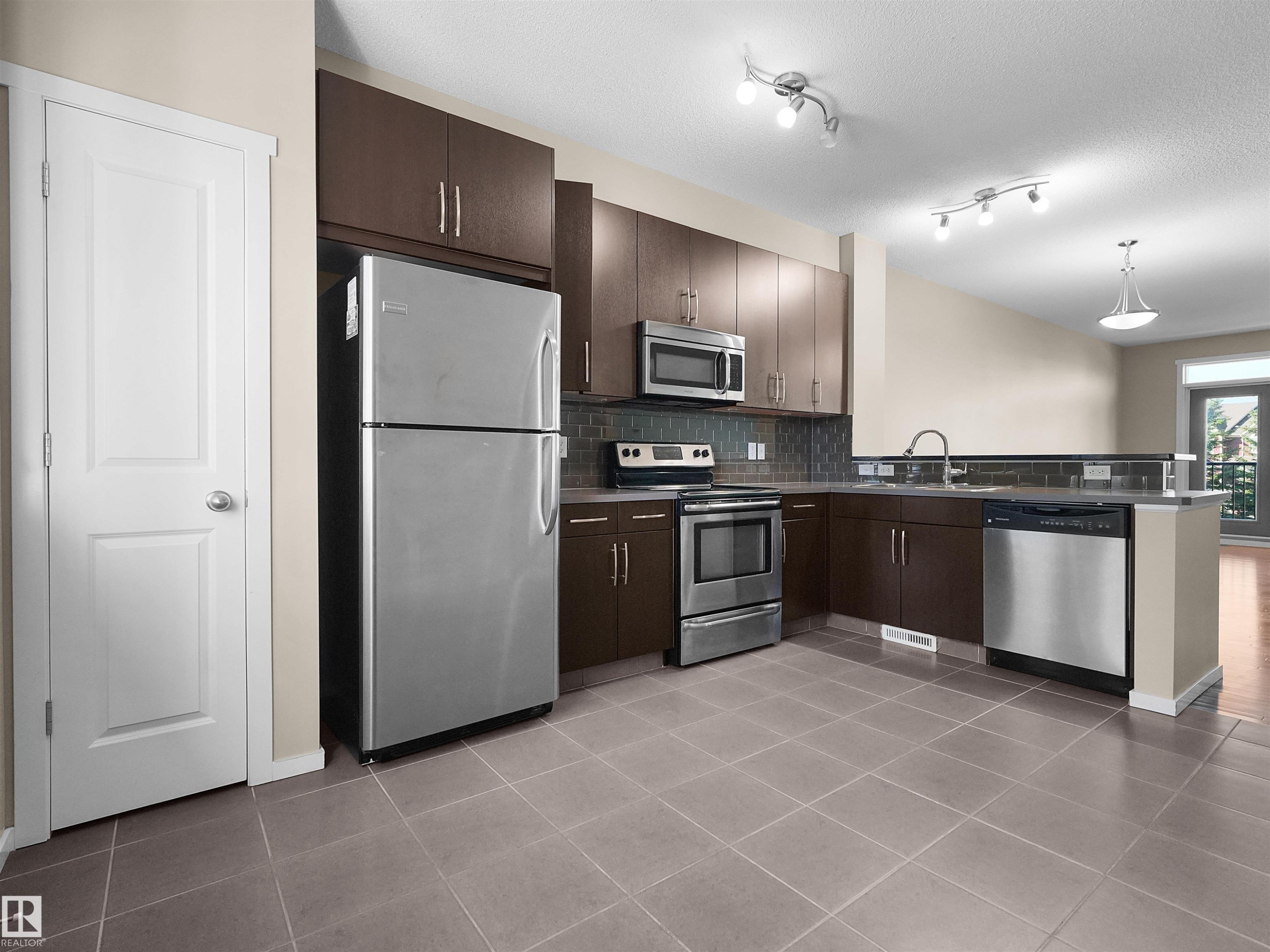 Kitchen featuring stainless steel appliances, dark wood-finish cabinetry, tile flooring, and track lighting - #162 603 Watt Bv Sw, Edmonton, AB