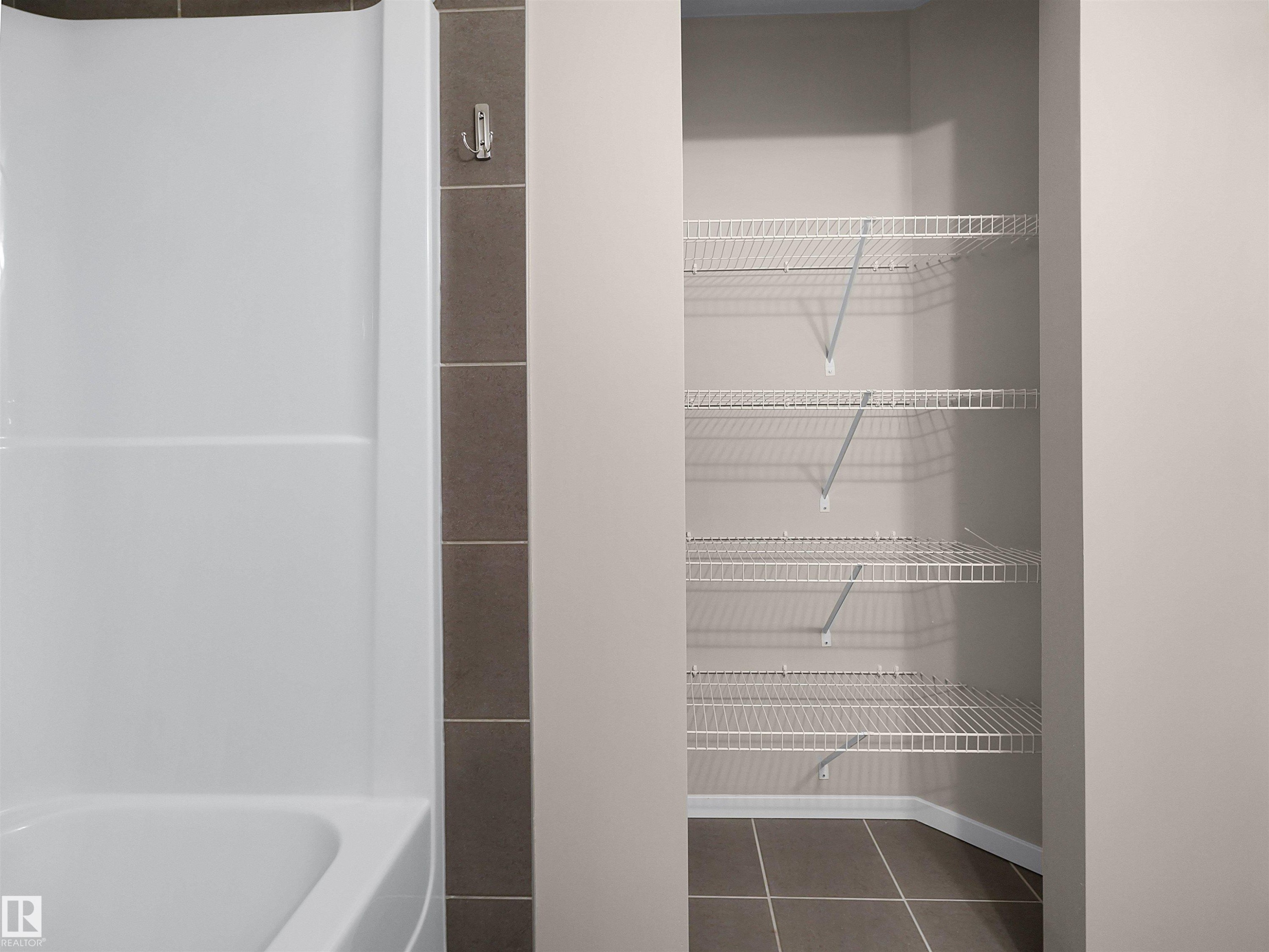 White bathtub with integrated shower, brown vertical tile accent, and a built-in closet with wire shelving and tile flooring - #162 603 Watt Bv Sw, Edmonton, AB