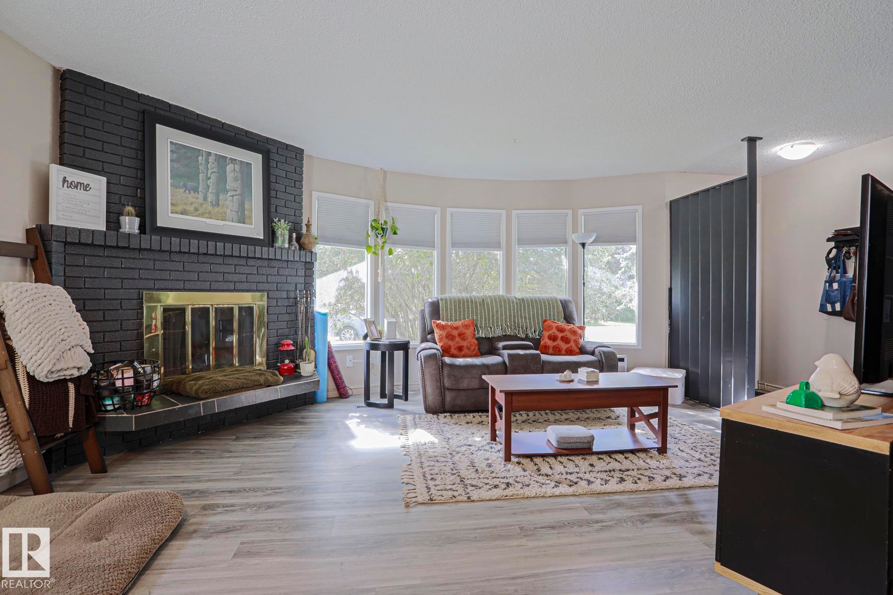 Black brick fireplace with brass-toned insert, wood-finish flooring, and a bay window - 21 Burlington Avenue, Spruce Grove, AB