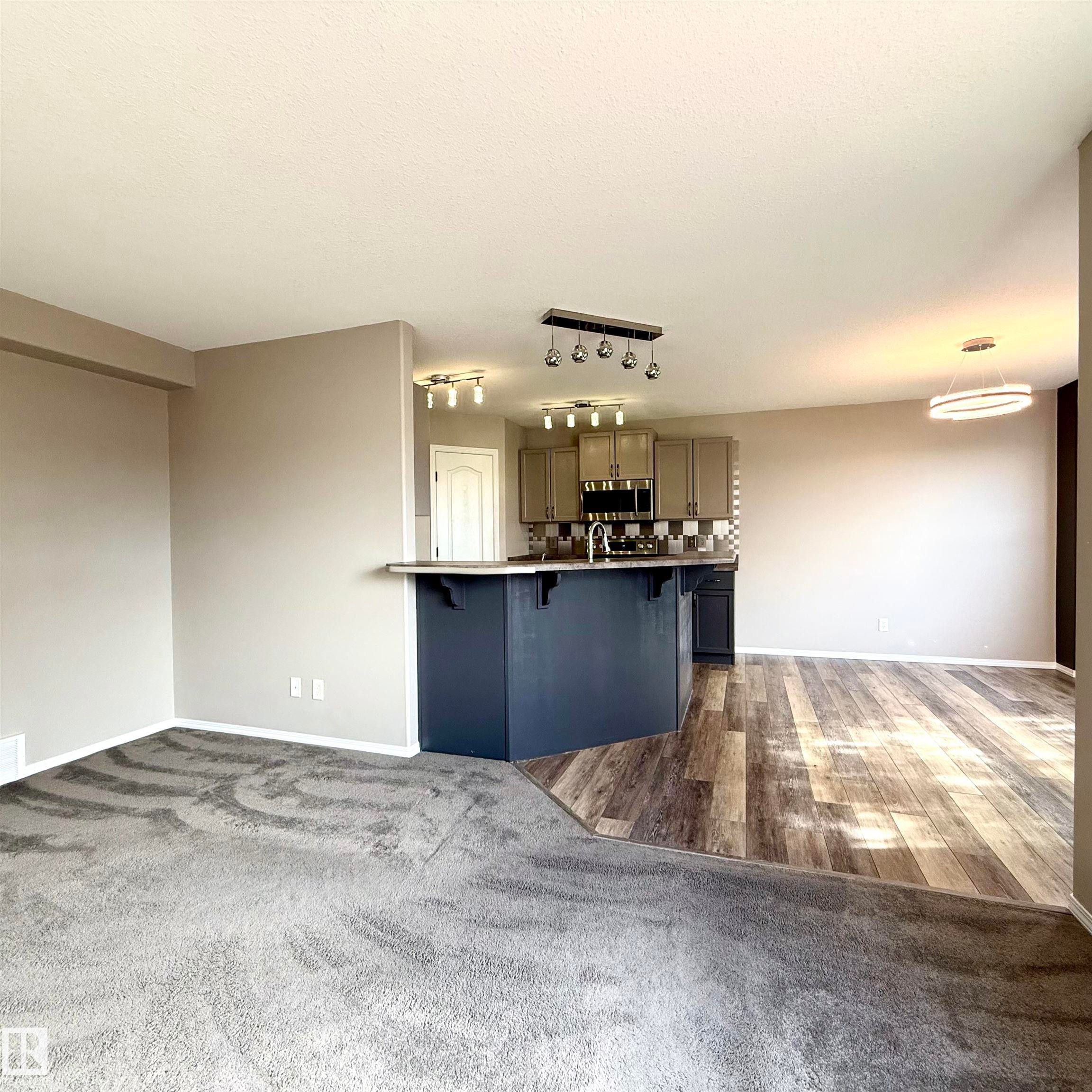 Open-concept living area with gray carpeting, wood-finish flooring, and a kitchen island with seating - 910 Oakland Boulevard, Devon, AB