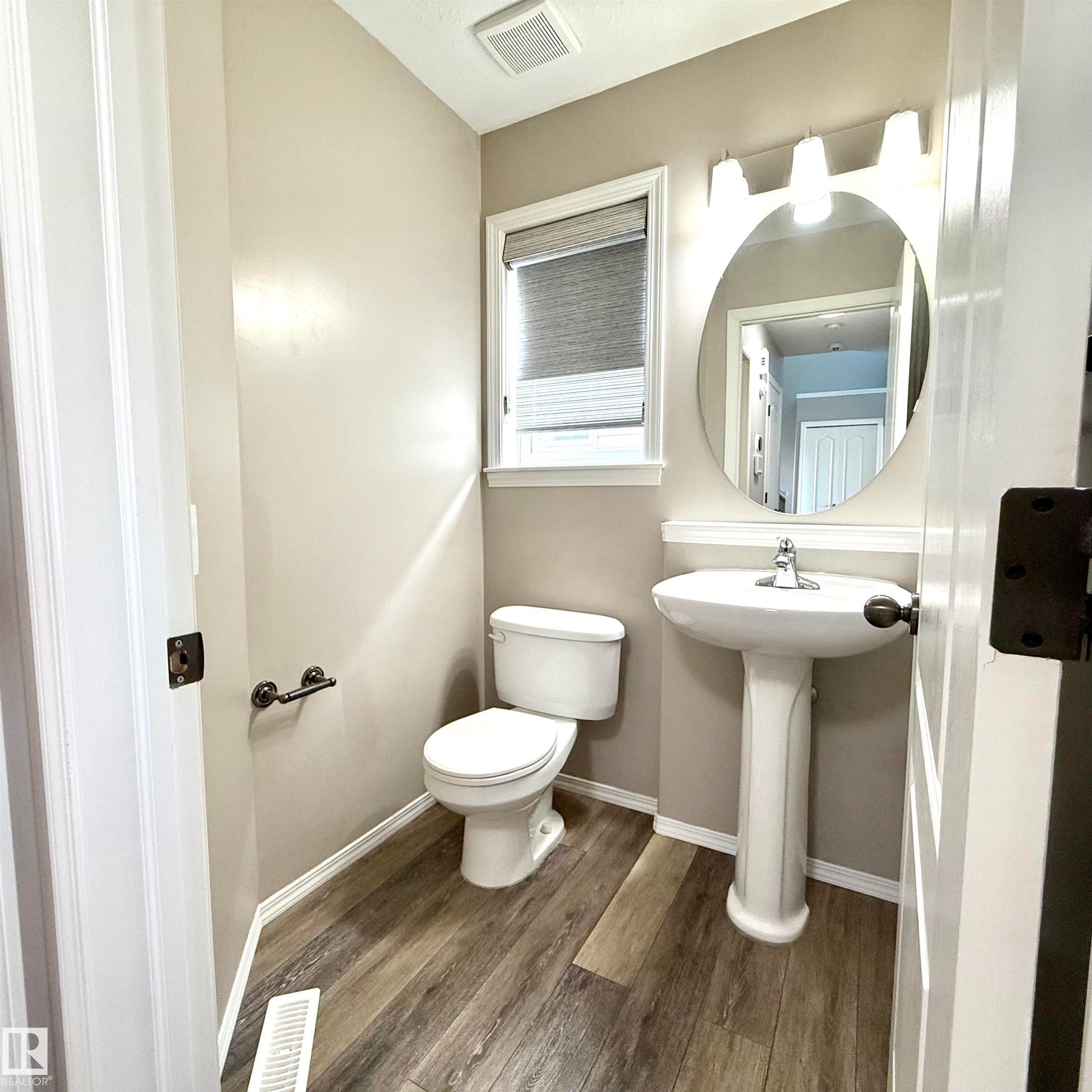 Wood-finish flooring, white pedestal sink, circular mirror, white window trim, and white toilet - 910 Oakland Boulevard, Devon, AB