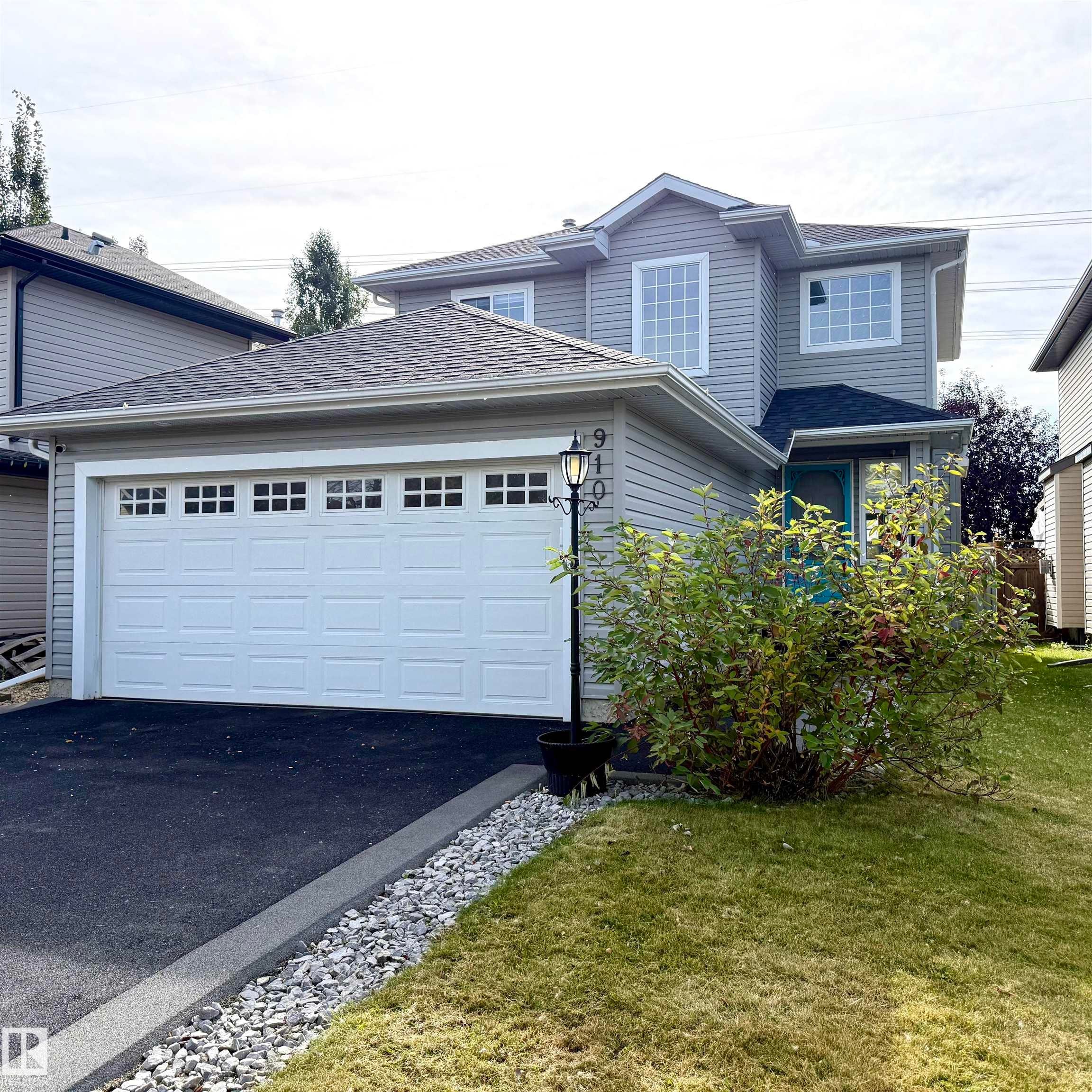 Two-story exterior with light gray siding, an attached garage featuring multiple window inserts, and an asphalt driveway - 910 Oakland Boulevard, Devon, AB