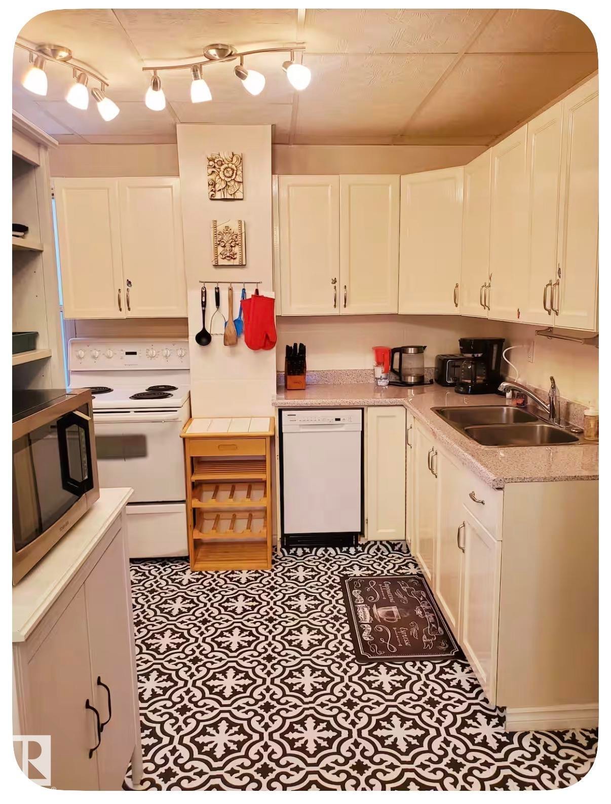 Kitchen featuring white cabinetry, track lighting, a double basin stainless steel sink, a white dishwasher, and patterned floor tile - 10649 125 Street Nw, Edmonton, AB