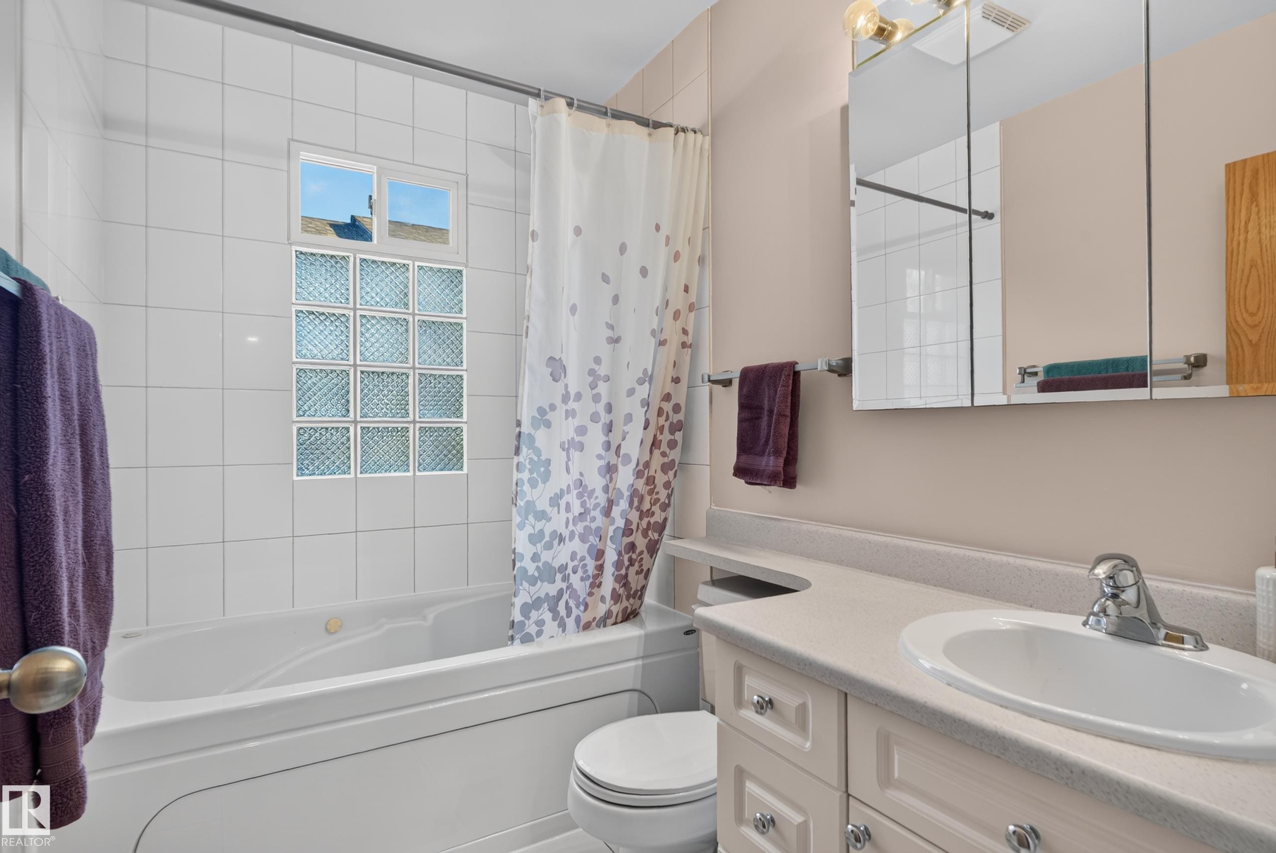 Built-in bathtub with tiled surround, glass block window, chrome faucet, vanity with integrated sink, and mirror with overhead lighting - 14611 109 Avenue Nw, Edmonton, AB