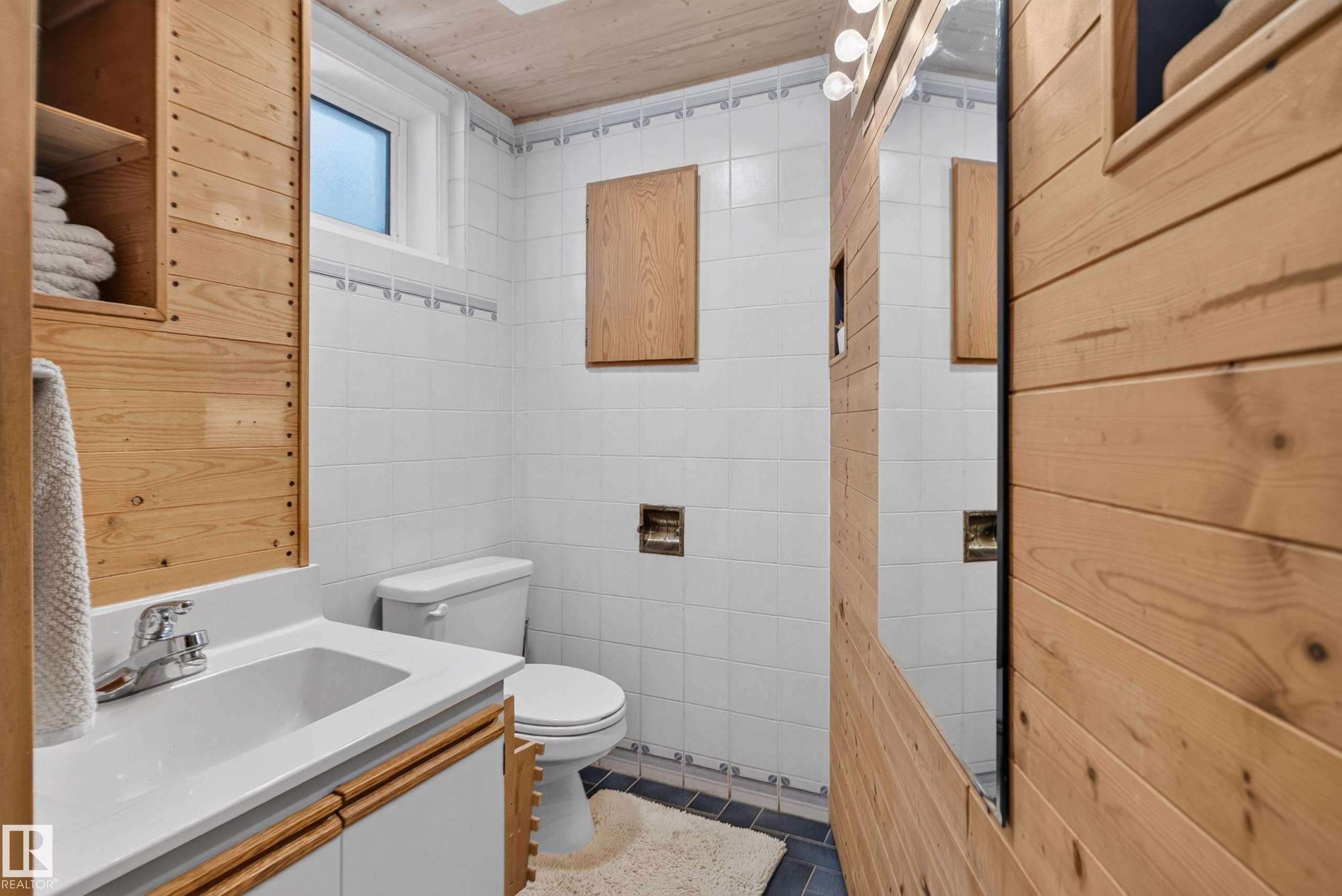 Wood-paneled walls and ceiling, white tiled walls with a decorative border, a white vanity with an integrated sink, and a mirrored wall panel with overhead lighting - 14611 109 Avenue Nw, Edmonton, AB