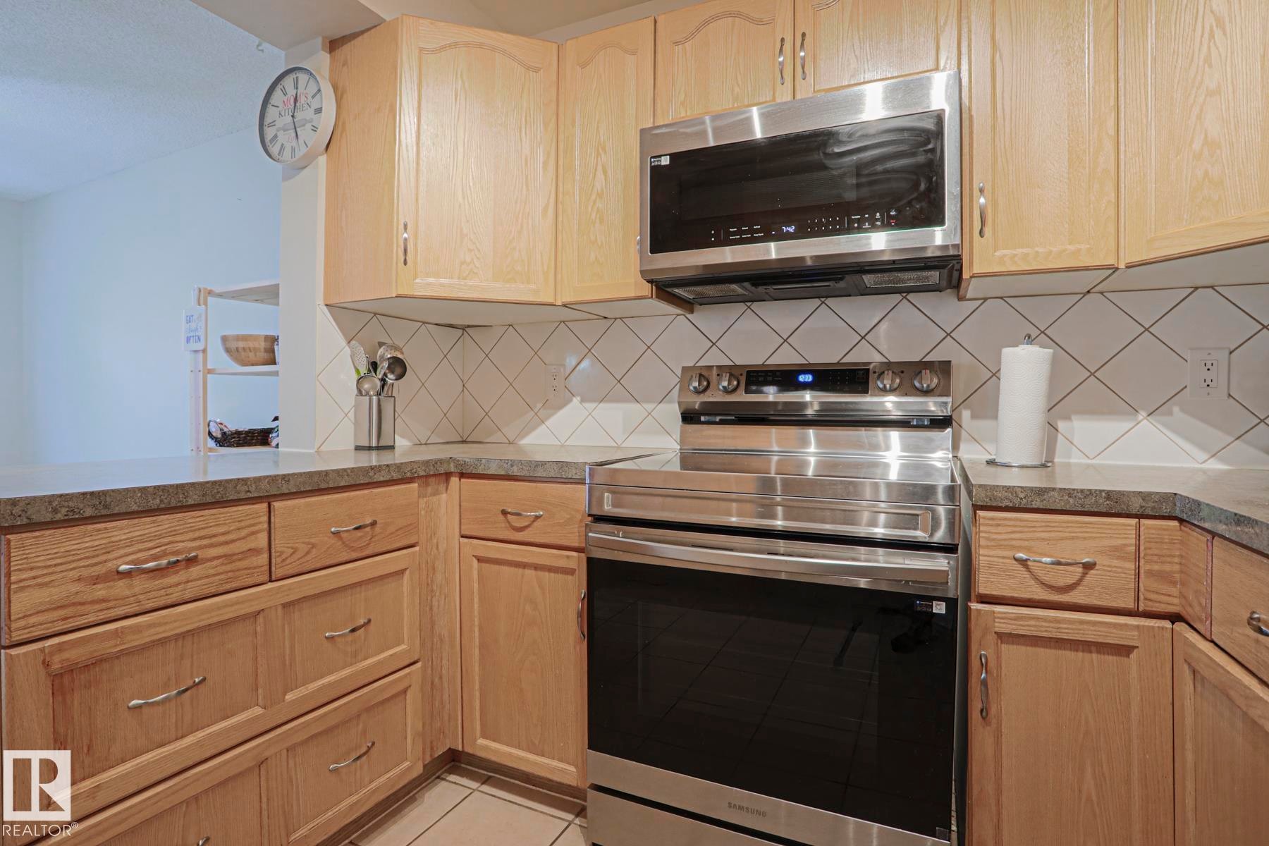 Wood-finish cabinetry, stainless steel oven, stainless steel microwave, stone-look countertops, and white tile backsplash - 319 Telford Court, Leduc, AB