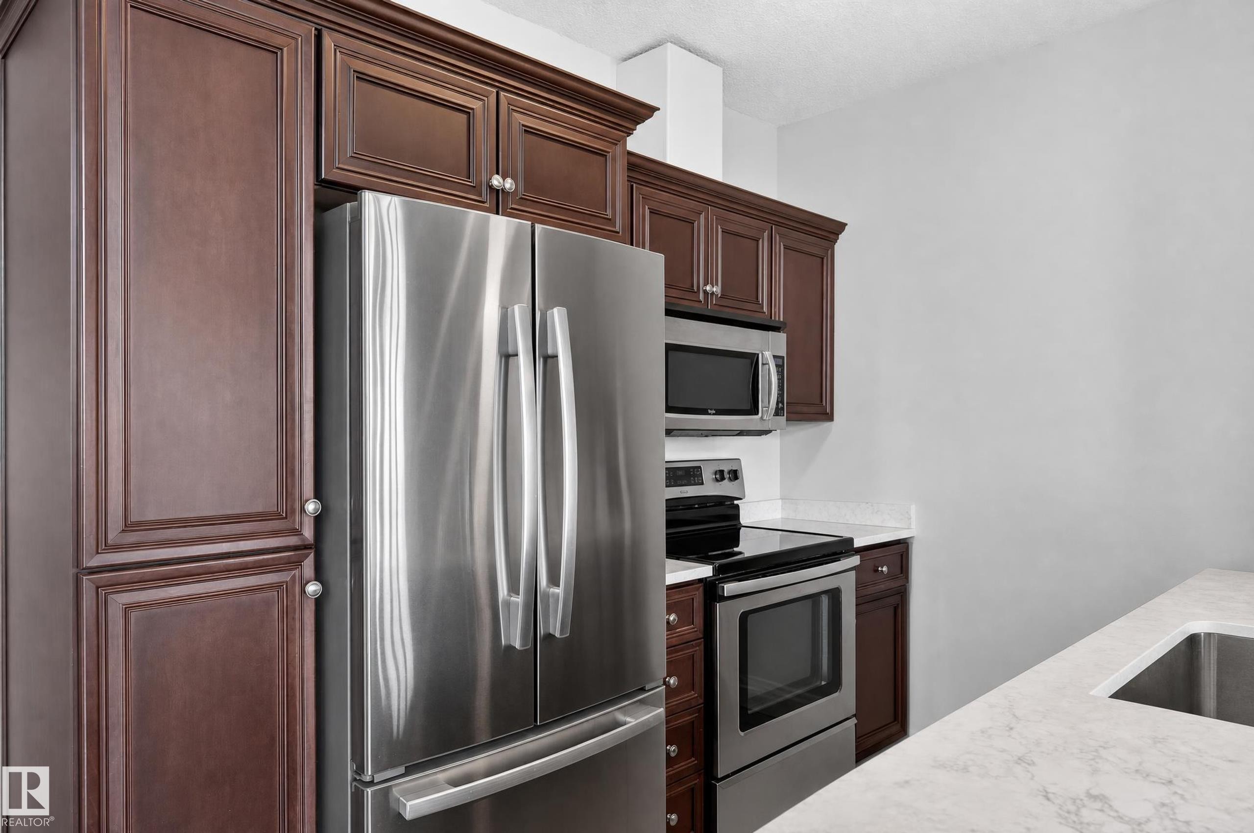 Stainless steel appliance package, dark wood cabinetry with crown molding, light-colored countertops, undermount sink, and recessed lighting - #411 304 Ambleside Link Li Sw, Edmonton, AB
