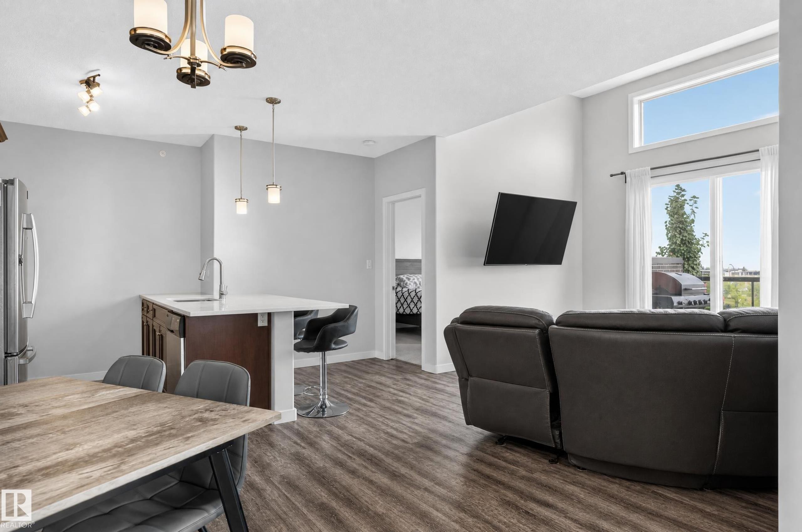 Open-concept living space featuring wood-finish flooring, a kitchen island with seating, stainless steel refrigerator, and large windows with a transom - #411 304 Ambleside Link Li Sw, Edmonton, AB