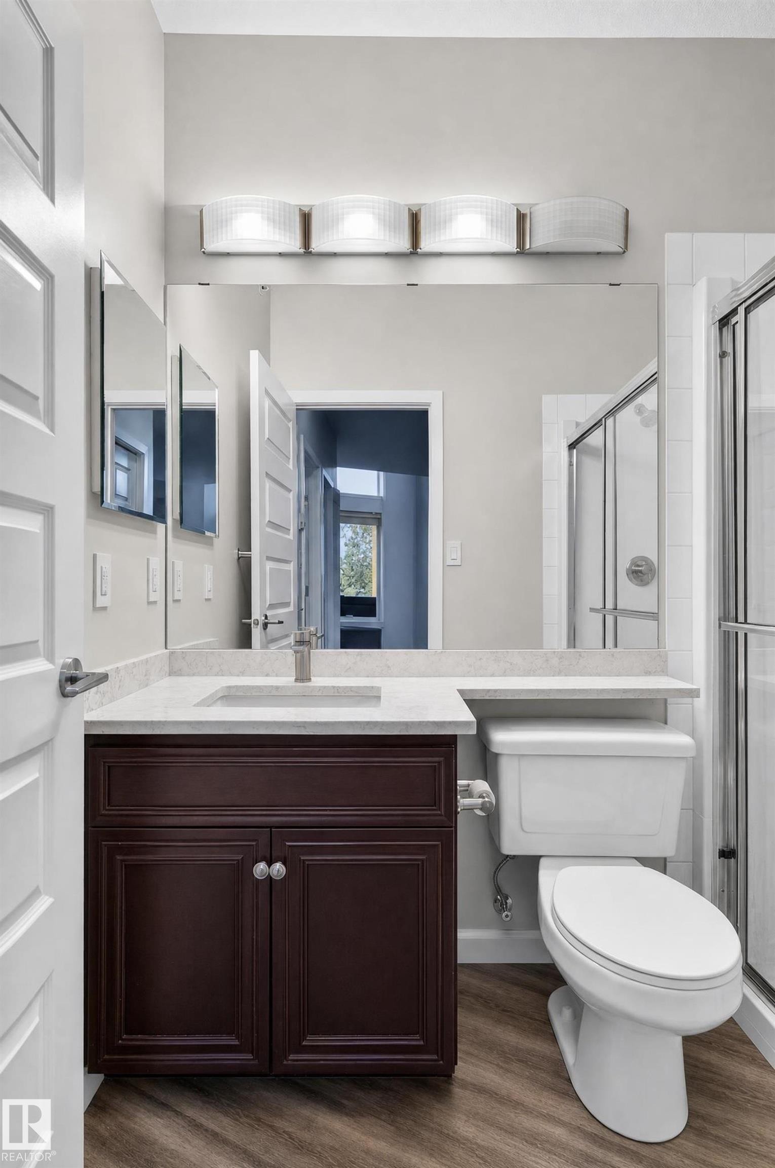Dark wood-finish vanity with a light-toned countertop, large wall-mounted mirror, brushed nickel faucet, and wood-finish flooring - #411 304 Ambleside Link Li Sw, Edmonton, AB