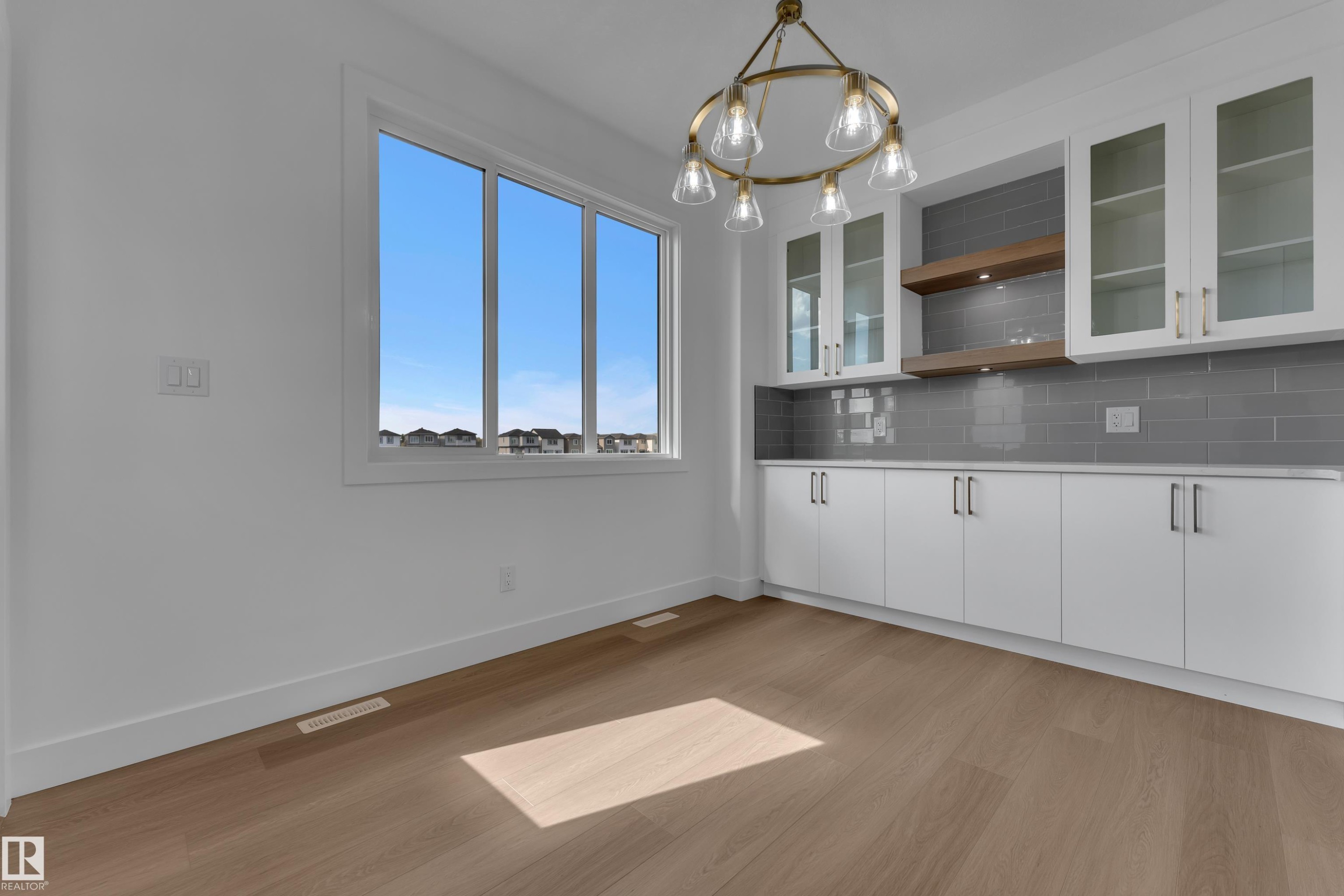 Wood-finish flooring with white baseboards, large multi-pane window, modern brass chandelier, built-in cabinetry with glass-front uppers, and gray subway tile backsplash - 4212 63 Street, Beaumont, AB