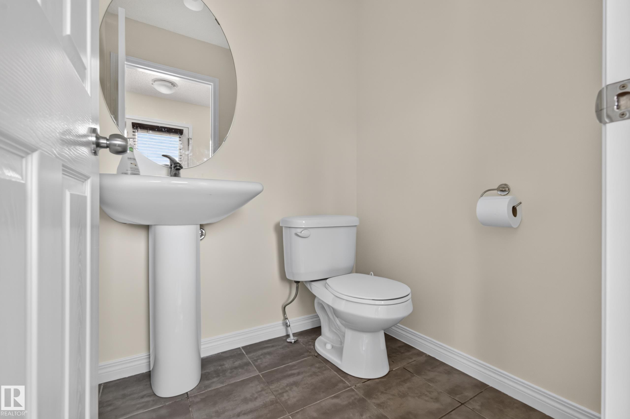Pedestal sink with a wall-mounted mirror, ceramic tile flooring, and a toilet with a brushed nickel toilet paper holder - 6933 21A Avenue Sw, Edmonton, AB