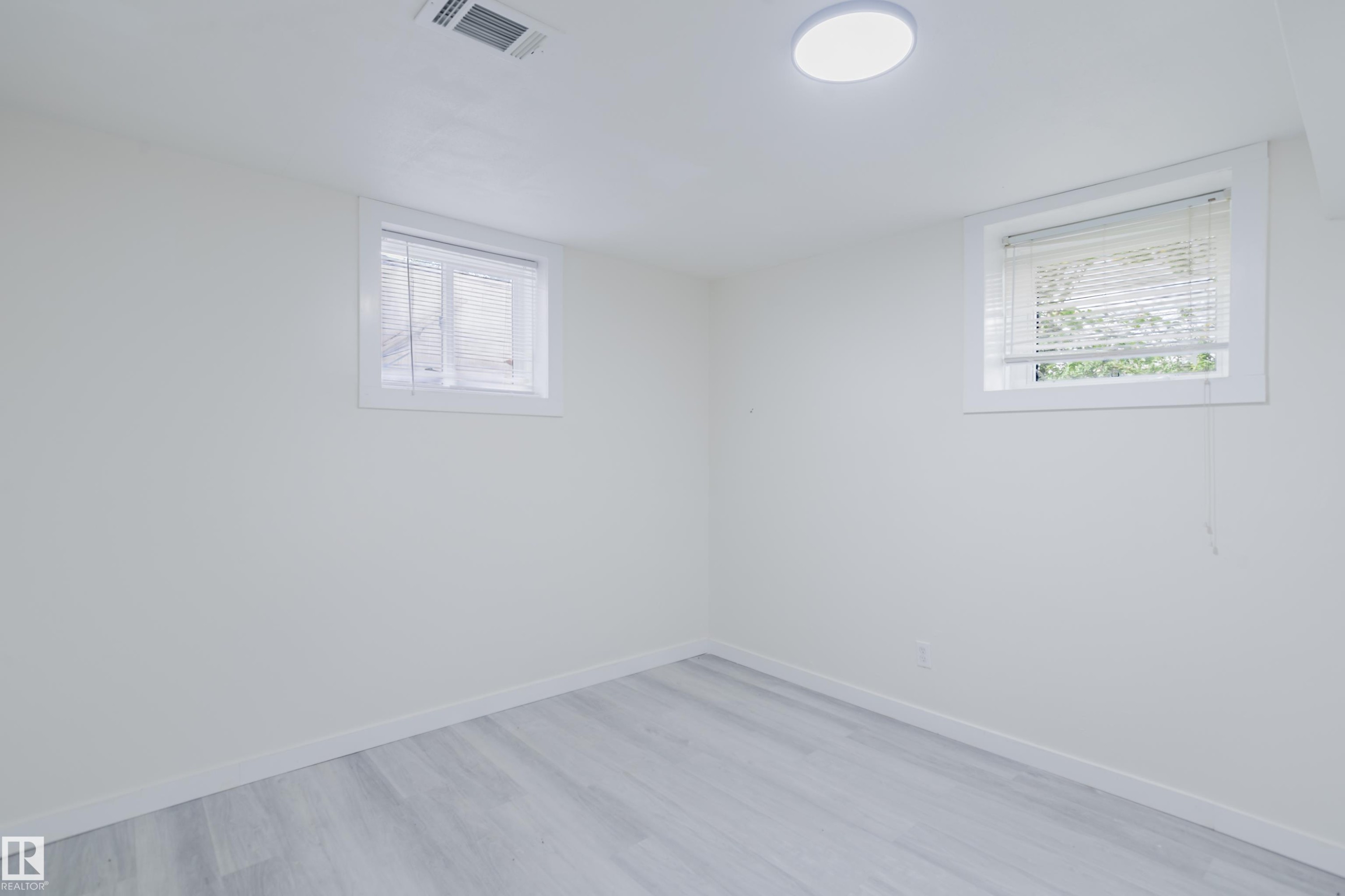 Light-toned wood-finish flooring, two rectangular windows with blinds, a white ceiling, and a flush-mount overhead light fixture - 11134 11138 116 Street Nw, Edmonton, AB