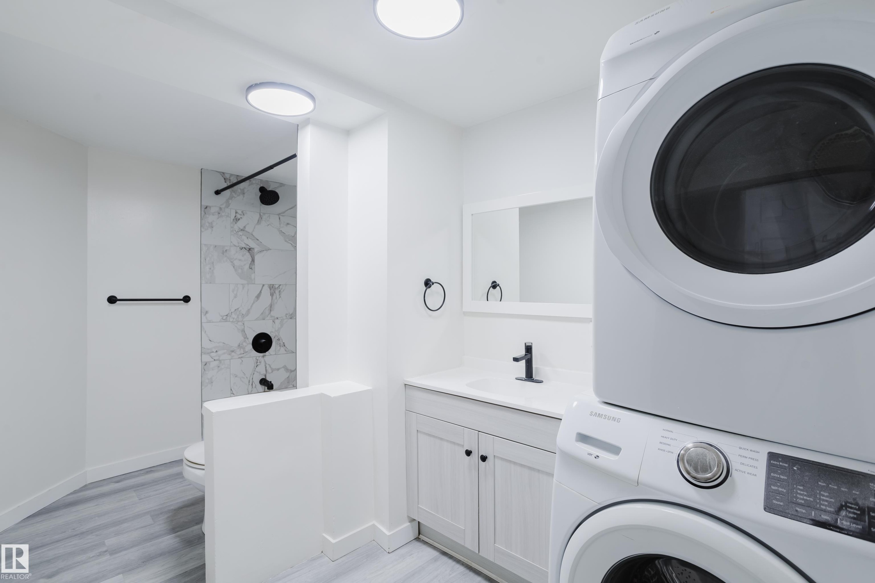 White stacked laundry appliances, vanity with a white countertop, wall-mounted mirror, and a shower with patterned wall tile - 11134 11138 116 Street Nw, Edmonton, AB