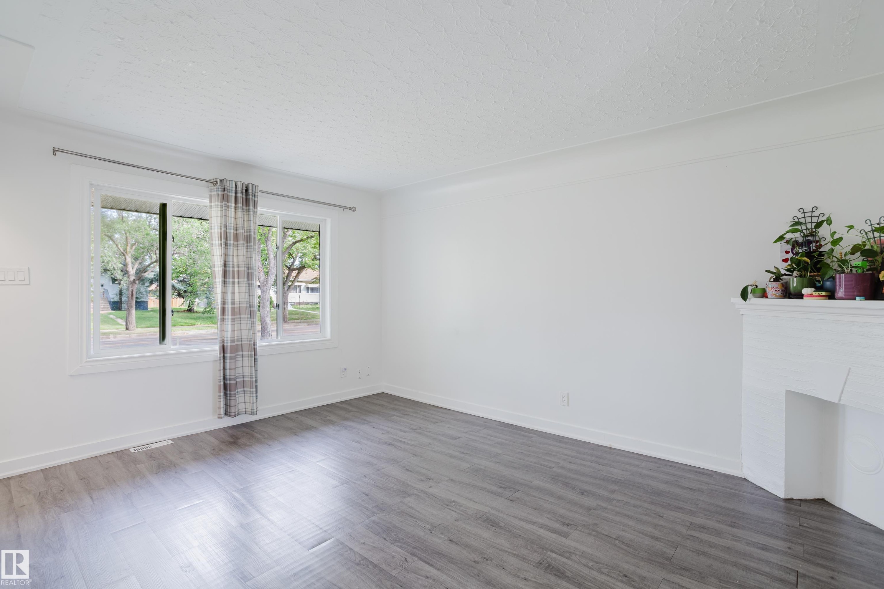 Spacious room featuring light gray wood-finish flooring, a white-painted fireplace, and a large window with white trim - 11134 11138 116 Street Nw, Edmonton, AB