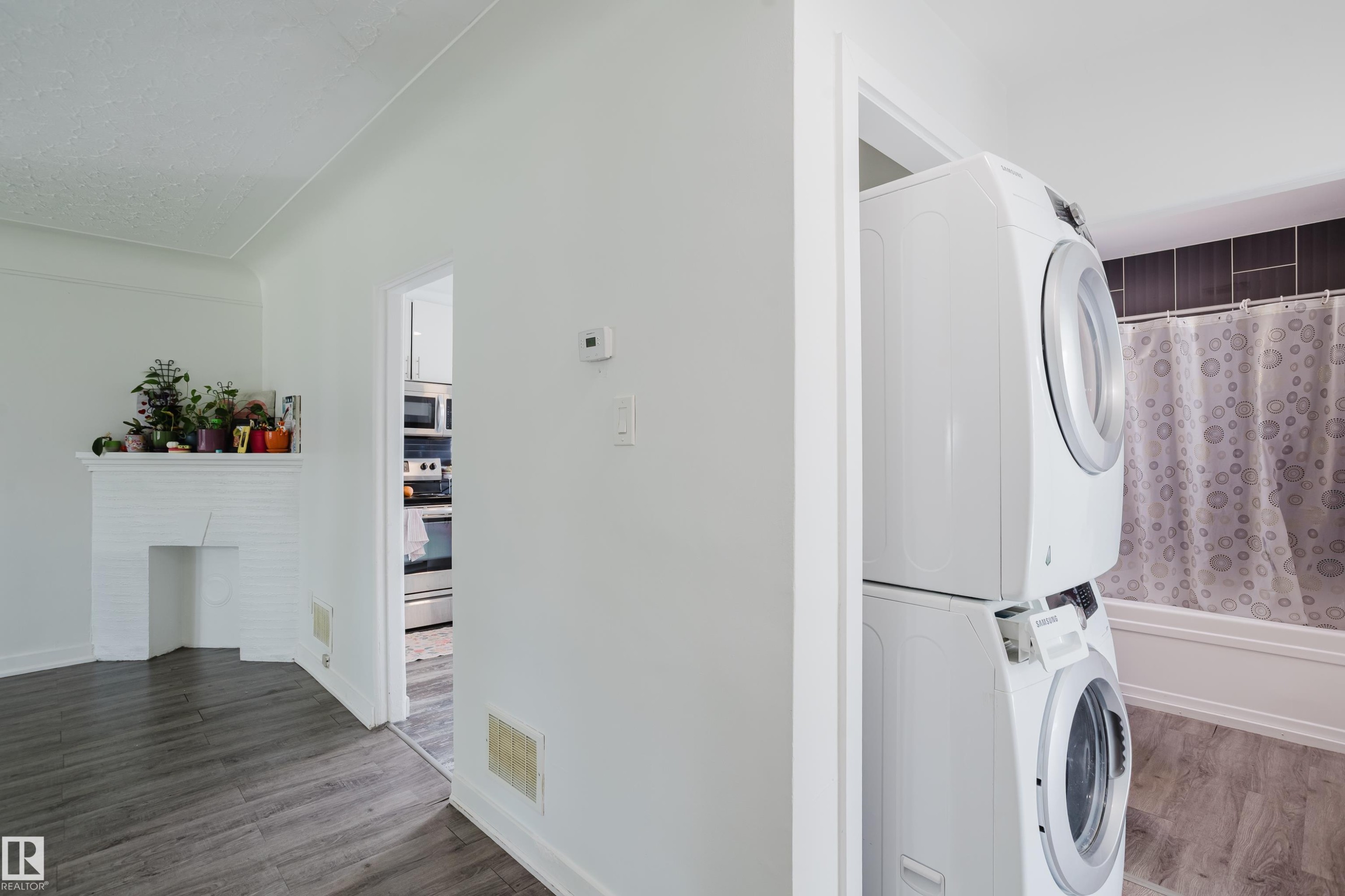 White brick fireplace, wood-finish flooring, white walls, stacked laundry appliances, and a full bathtub with dark tile surround - 11134 11138 116 Street Nw, Edmonton, AB
