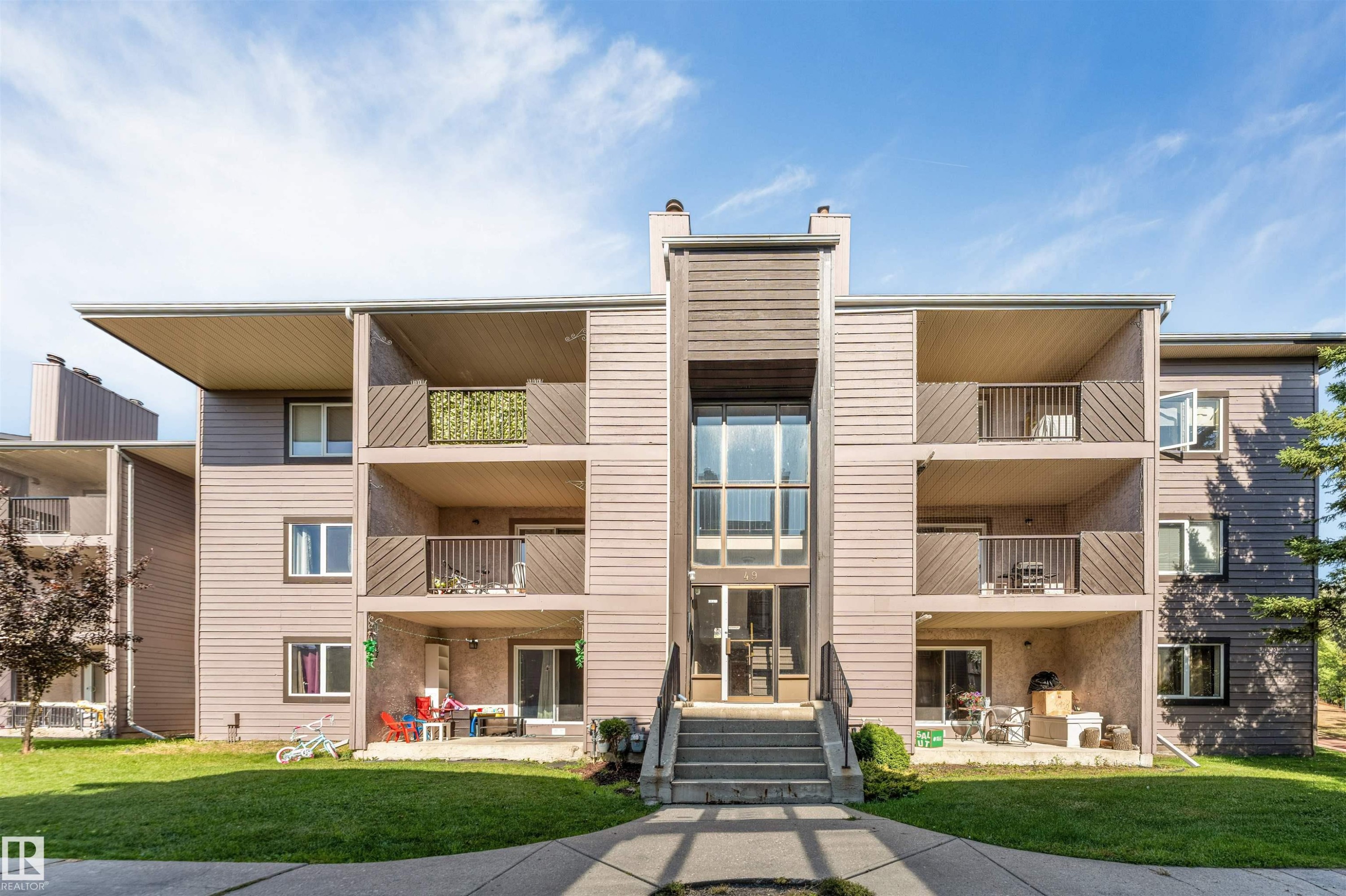 Multi-story building with horizontal siding, multiple balconies with metal railings, a central glass-enclosed entry, and an exterior staircase - #101 49 Akins Dr, St. Albert, AB