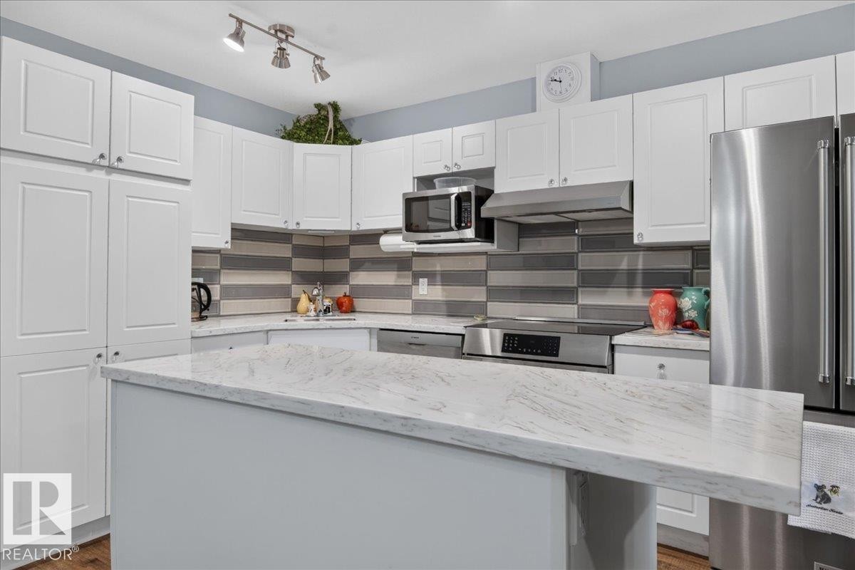 White shaker-style cabinetry, stainless steel appliances, patterned tile backsplash, track lighting, and a kitchen island with a light-colored countertop - #125 4610 50 Ave, Stony Plain, AB