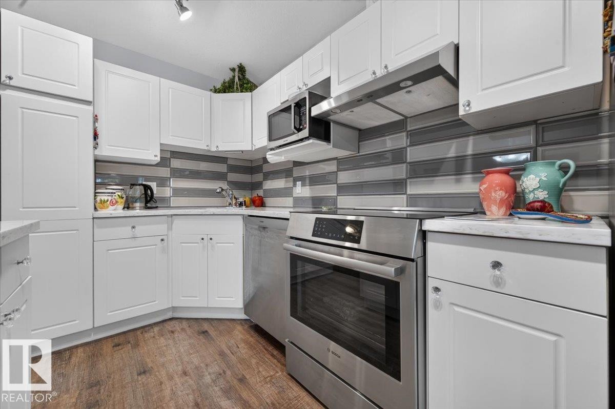White shaker-style cabinetry, stainless steel oven, built-in microwave, wood-finish flooring, and a glass subway tile backsplash - #125 4610 50 Ave, Stony Plain, AB