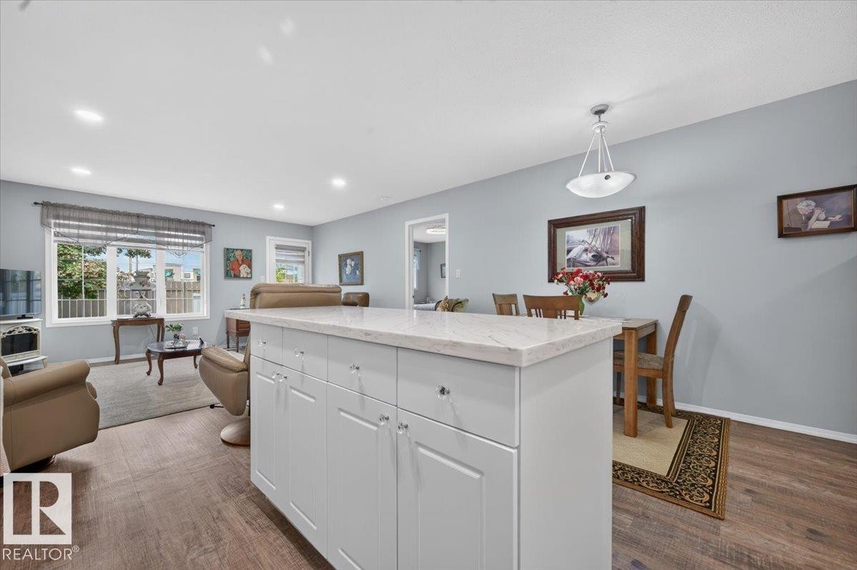 Open-concept living space featuring wood-finish flooring, recessed lighting, a kitchen island with white cabinetry and a light-toned countertop, and a dining area with a pendant light fixture - #125 4610 50 Ave, Stony Plain, AB