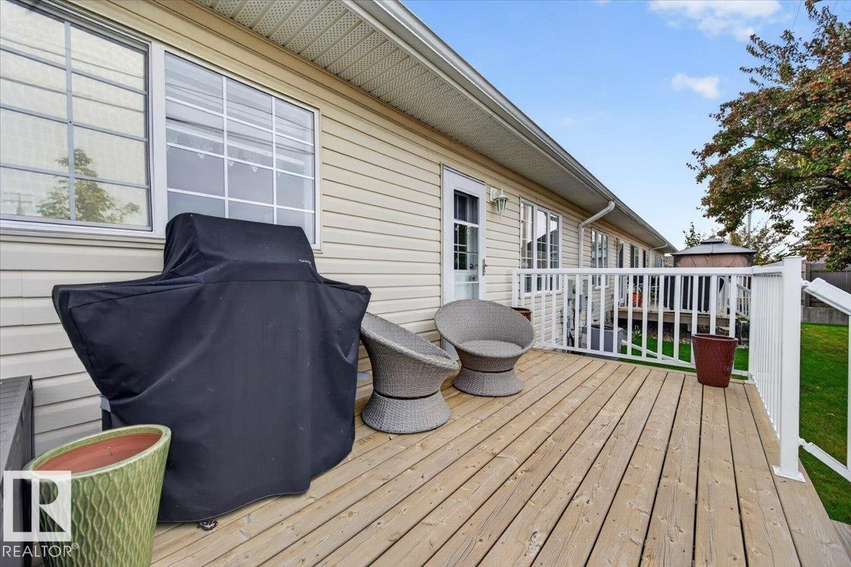 Wood-plank deck with white railing, featuring exterior siding and a clear glass paneled door - #125 4610 50 Ave, Stony Plain, AB