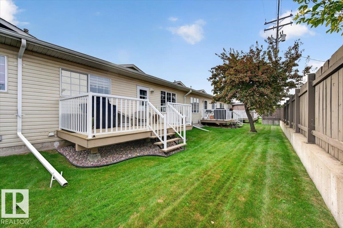 Expansive green space featuring a wood deck with white railings, mature tree, and a wood privacy fence on a concrete retaining wall - #125 4610 50 Ave, Stony Plain, AB