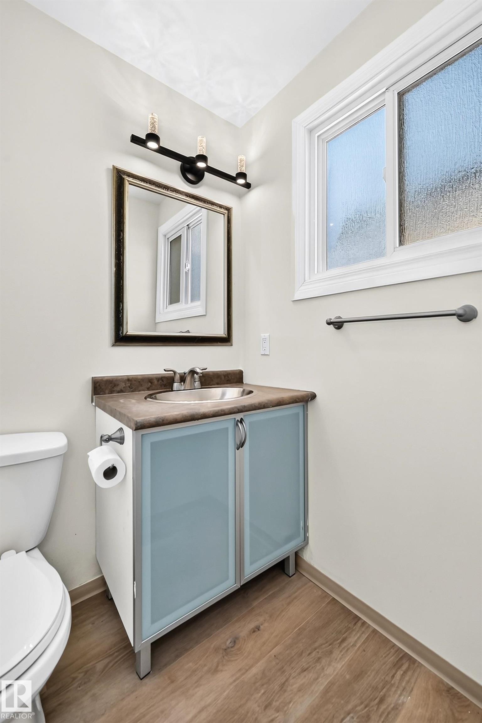 Wood-finish flooring, vanity with frosted cabinet doors, countertop with integral sink, framed mirror, and a three-light fixture - 161 Lancaster Terrace Nw, Edmonton, AB