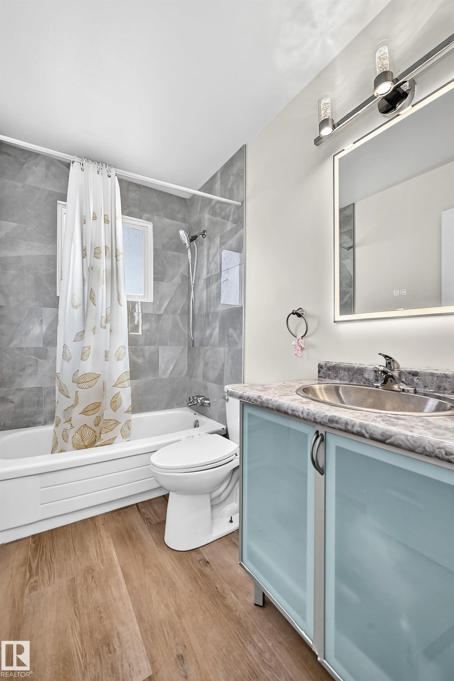 Wood-finish flooring, full bathtub with shower, gray tile surround, light blue vanity with frosted glass doors, and a back-lit mirror - 161 Lancaster Terrace Nw, Edmonton, AB