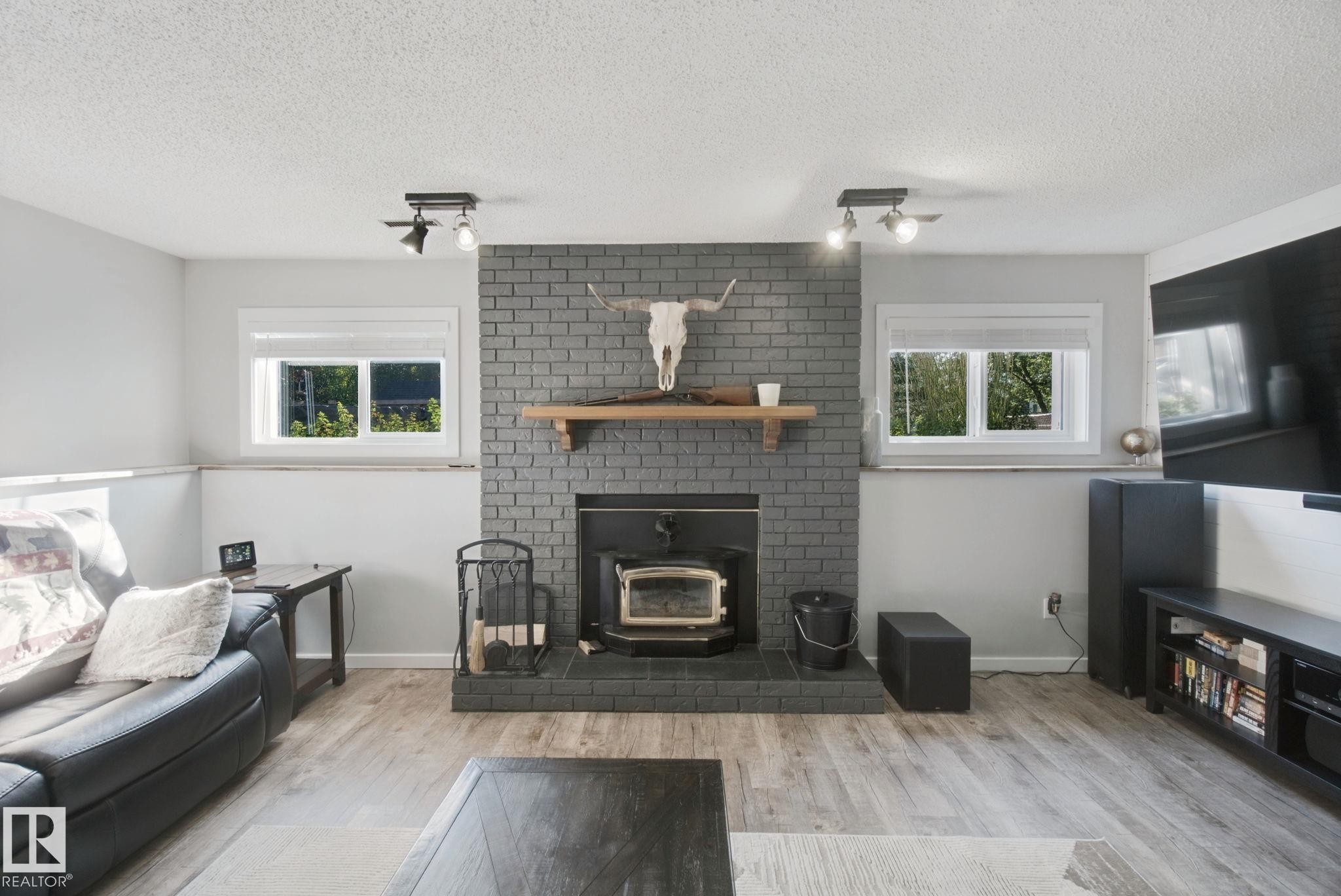 Brick fireplace with wood-burning stove, wood-finish flooring, recessed windows, and track lighting fixtures - 38 Menalta Place, Cardiff, AB
