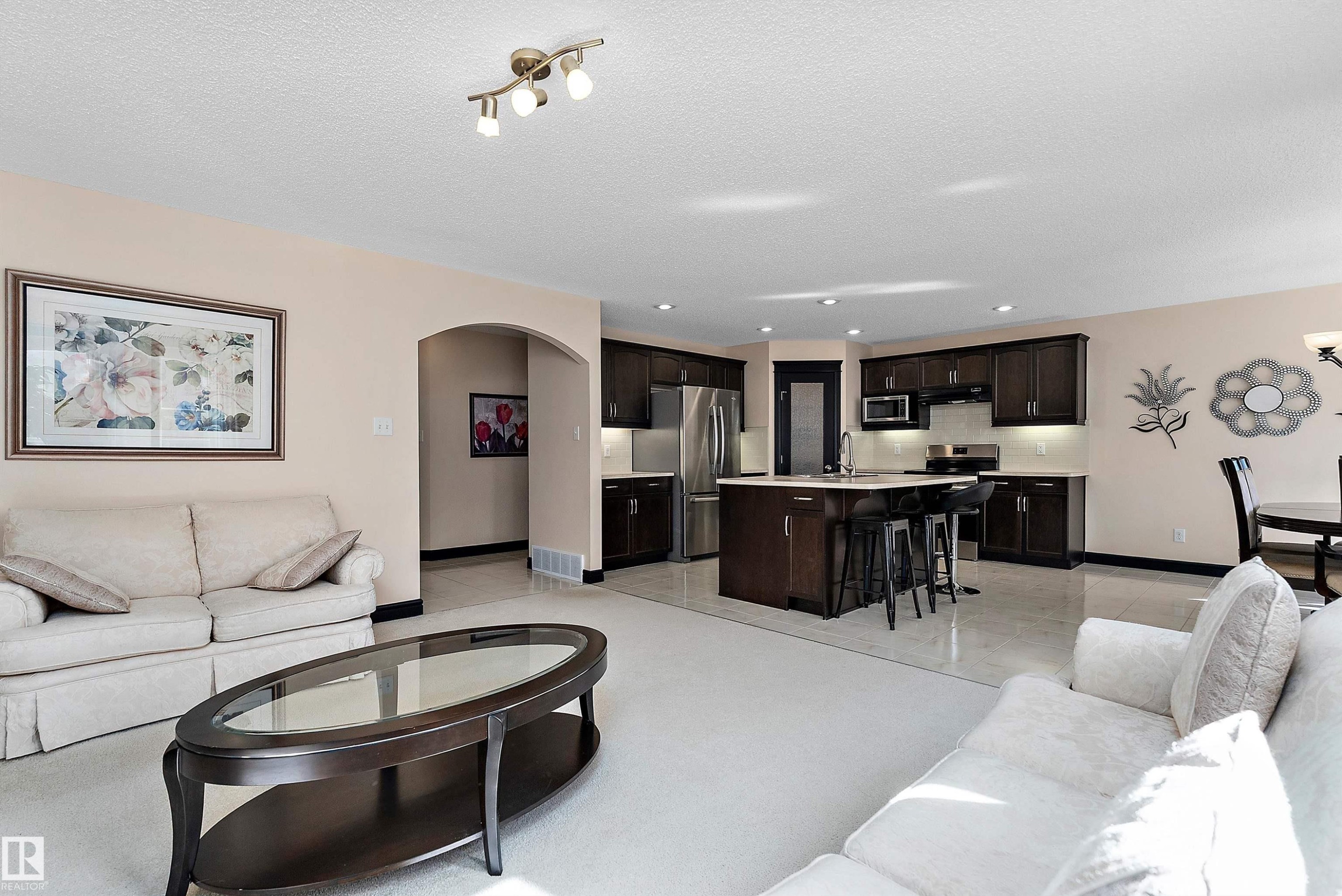 Open-concept living area with carpet flooring, arched doorway, kitchen island with seating, stainless steel appliances, and track lighting - 11515 16 Avenue Sw, Edmonton, AB