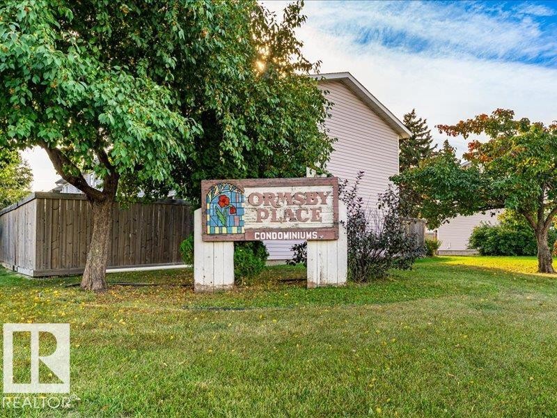 Property entrance featuring a "Ormsby Place Condominiums" sign, mature trees, and a wooden fence - 18325 66 Avenue Nw, Edmonton, AB