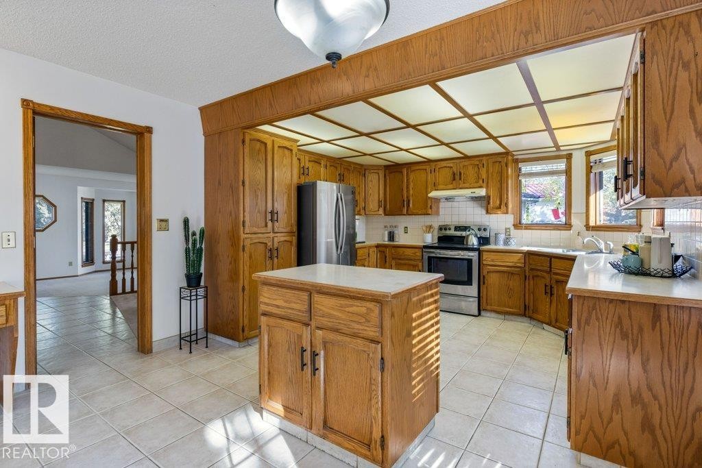 Kitchen featuring wood cabinetry, white tile flooring, a center island with countertop, stainless steel appliances, and a coffered ceiling - 507 Burton Close Nw, Edmonton, AB