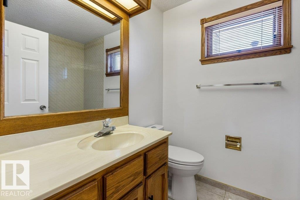 Bathroom featuring a wood-framed vanity mirror, built-in vanity cabinet with integrated sink, window with wood trim, white toilet, and tiled flooring - 507 Burton Close Nw, Edmonton, AB
