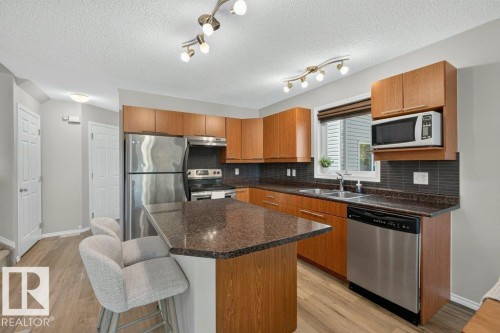 Kitchen featuring wood-finish cabinetry, stainless steel appliances, dark stone-finish countertops, and a tile backsplash - #41 287 Macewan Rd Sw, Edmonton, AB 