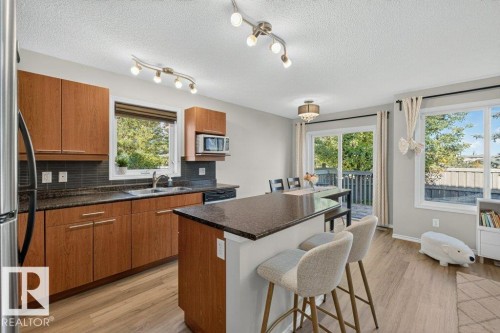 Wood-finish flooring throughout, wood cabinetry, dark countertops, dark tile backsplash, and a sliding glass door to an outdoor deck area - #41 287 Macewan Rd Sw, Edmonton, AB 