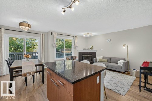 Wood-finish flooring throughout, kitchen island with dark stone countertop, built-in fireplace with dark tile surround, track lighting, and a sliding glass door opening to a deck - #41 287 Macewan Rd Sw, Edmonton, AB 