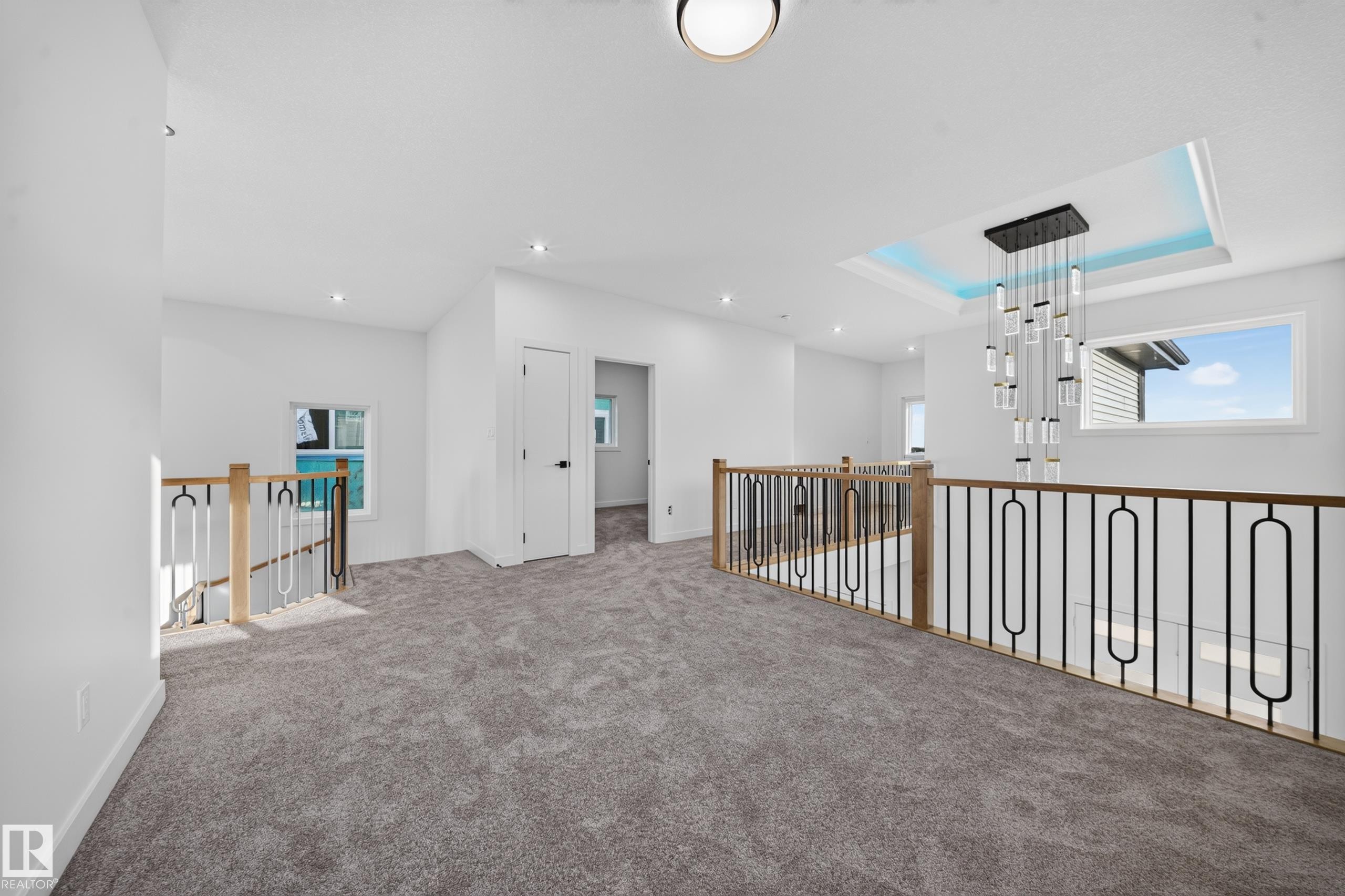 Carpeted hallway featuring a recessed ceiling fixture, contemporary chandelier, and decorative metal railings with wood accents - 13024 186 Avene Nw, Edmonton, AB
