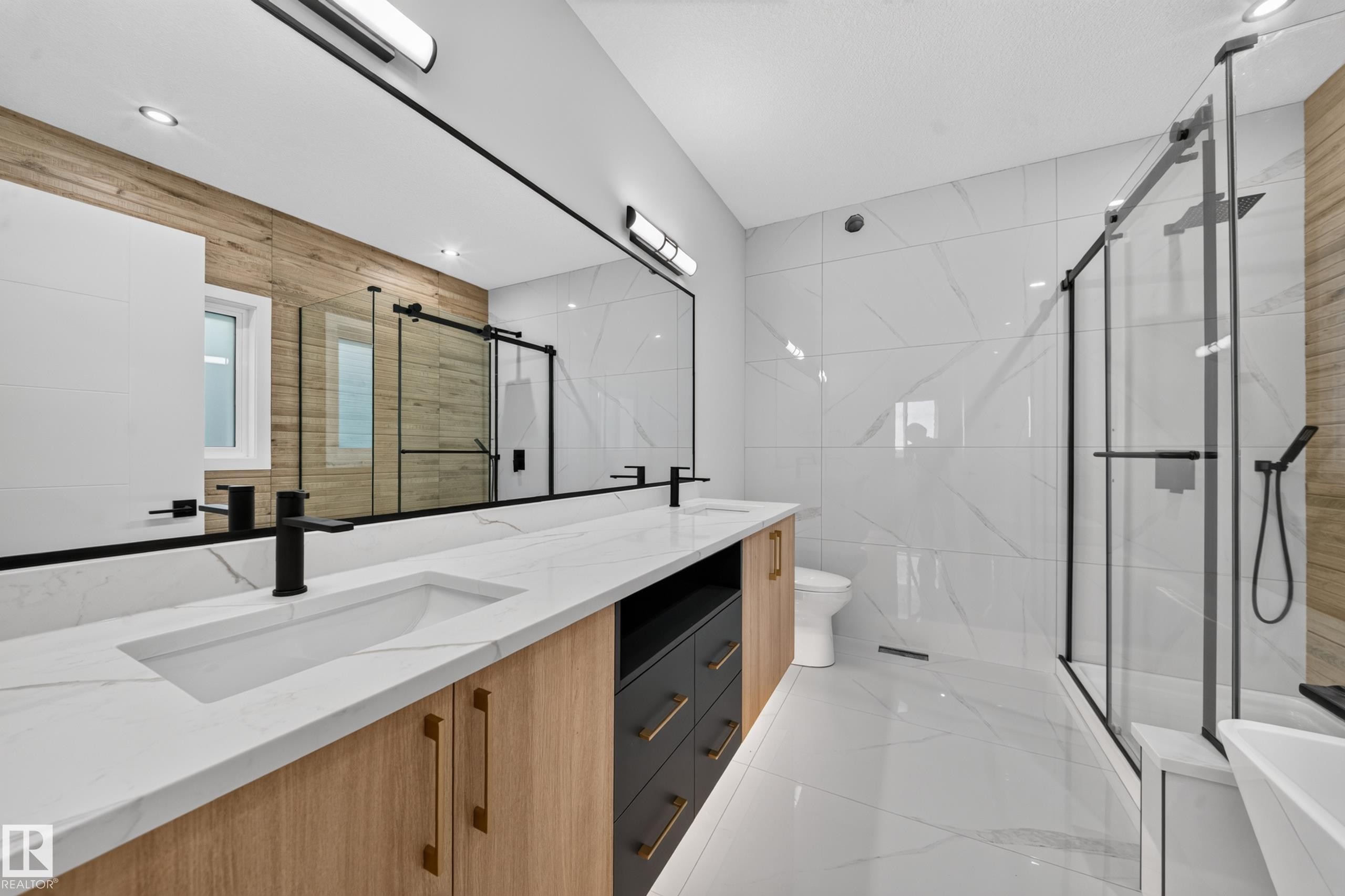 Dual vanity with integrated sinks and black matte faucets, expansive mirror with task lighting, wood-finish and dark cabinetry, large format wall and floor tiling, and a frameless glass shower enclosure - 13024 186 Avene Nw, Edmonton, AB