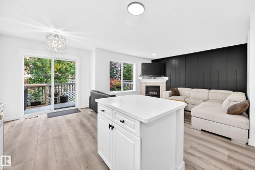 Open-concept living area with wood-finish flooring, a kitchen island with white cabinetry, a fireplace, and a sliding glass door opening to an outdoor deck - #27 287 Macewan Rd Sw, Edmonton, AB 