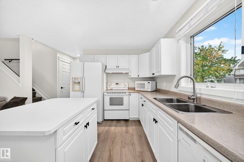Kitchen featuring white cabinetry, a central island, wood-finish flooring, a double basin sink, and a large window - #27 287 Macewan Rd Sw, Edmonton, AB 