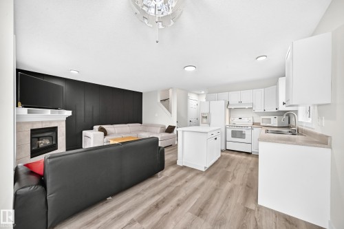 Open-concept living space featuring wood-finish flooring, a kitchen island, white cabinetry, a tiled fireplace with a mantel, and recessed lighting - #27 287 Macewan Rd Sw, Edmonton, AB 