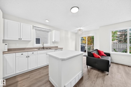 White kitchen cabinetry, wood-finish flooring, central kitchen island, sliding glass patio door, and large window - #27 287 Macewan Rd Sw, Edmonton, AB 
