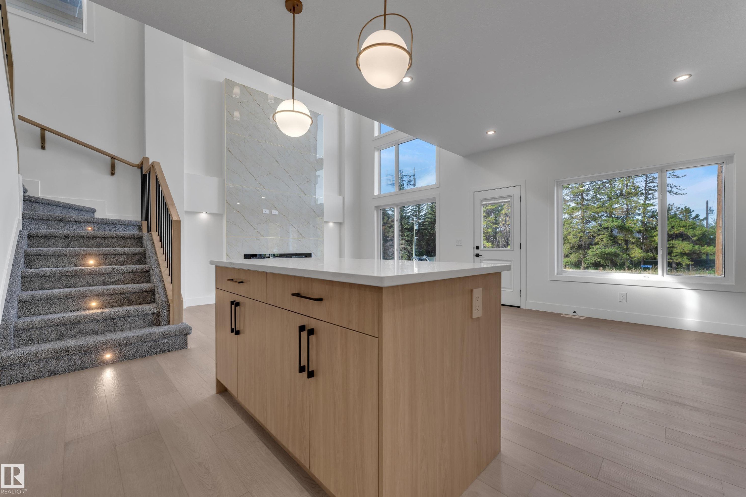 Open-concept room featuring light wood-finish flooring, a contemporary kitchen island with a light countertop, a staircase with carpeted treads and wood handrail, and high ceilings with recessed lighting - 247 Linden Loop, Leduc, AB