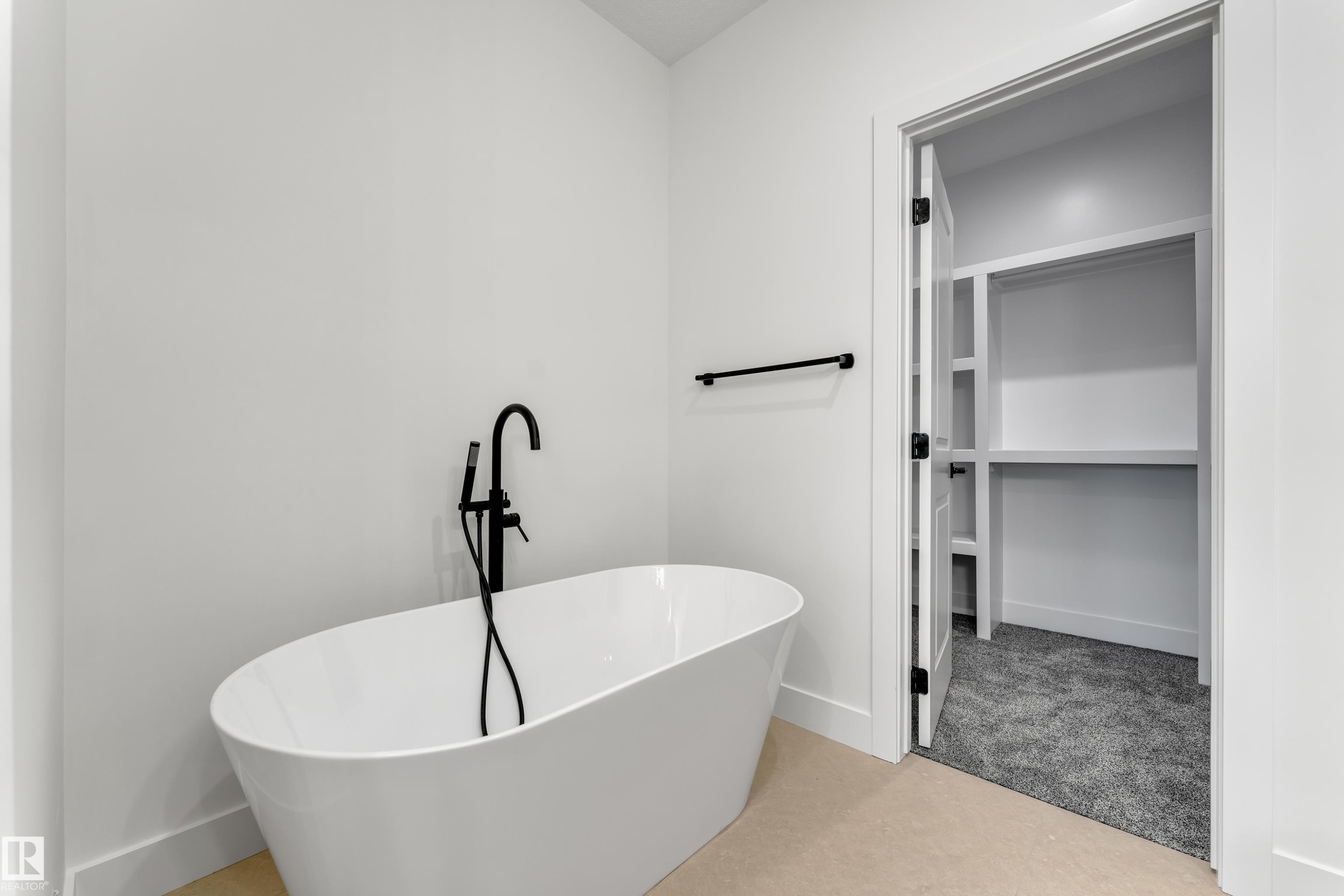 Freestanding soaking tub with matte black floor-mounted faucet, light-toned flooring, and an open closet with built-in shelving and gray carpeting - 247 Linden Loop, Leduc, AB